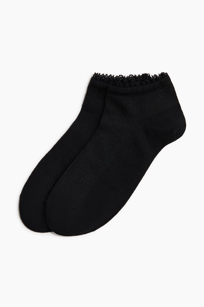 A pair of black ankle socks is lying flat, one subtly overlapping the other. The socks are featuring a delicate lace trim along the top edge and a fine ribbed texture throughout the main body. - 1