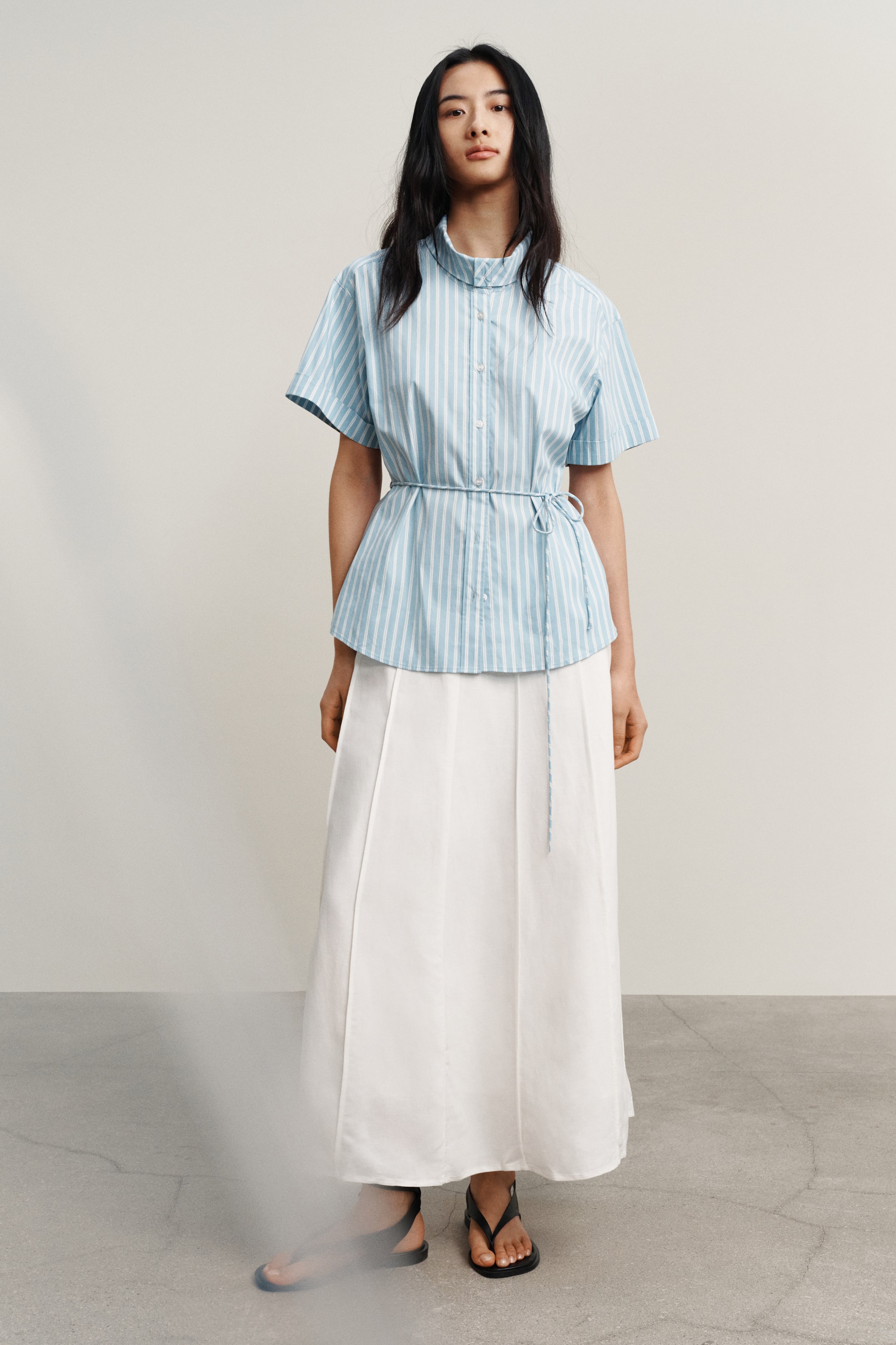 View larger image: A set pakaian sedang dipersembahkan, menampilkan kemeja berjalur menegak biru muda dan putih dengan lengan pendek, kolar band, dan hadapan berkancing. Kemeja ini diikat di pinggang dengan tali pinggang biru nipis, melabuh di atas skirt maxi putih dengan garis jahitan menegak, dan dilengkapkan dengan sandal rata hitam.
