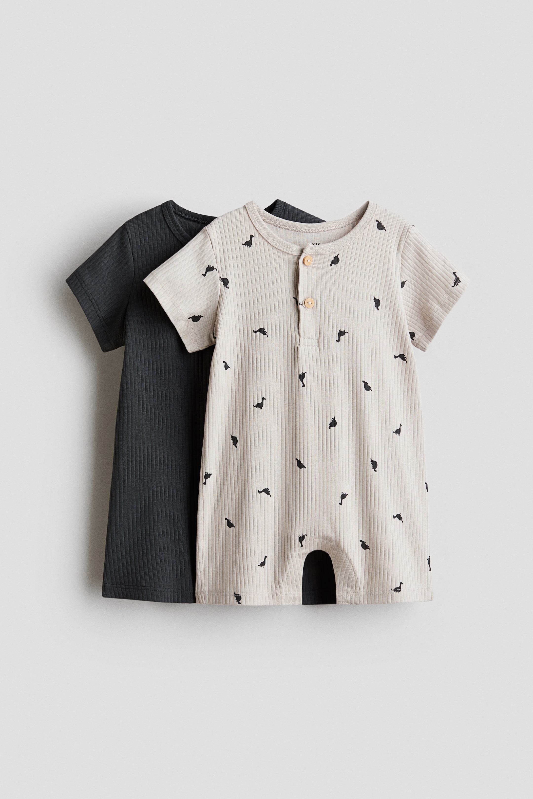 View larger image: A light beige baby romper, facing FRONT, showcases a ribbed texture, short sleeves, and short leg openings, adorned with a pattern of small dark dinosaur silhouettes and a two-button Henley placket. Behind it, a dark grey baby romper is visible, also highlighting a ribbed texture and short sleeves.