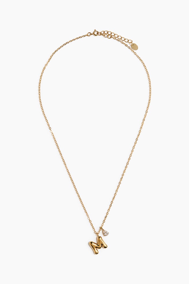 A gold-toned necklace is showcasing a delicate cable chain with a three-dimensional, puffy capital letter "M" pendant and a small, teardrop-shaped clear gemstone charm. Facing front, the necklace features an adjustable clasp with an extender chain at the top. - 1
