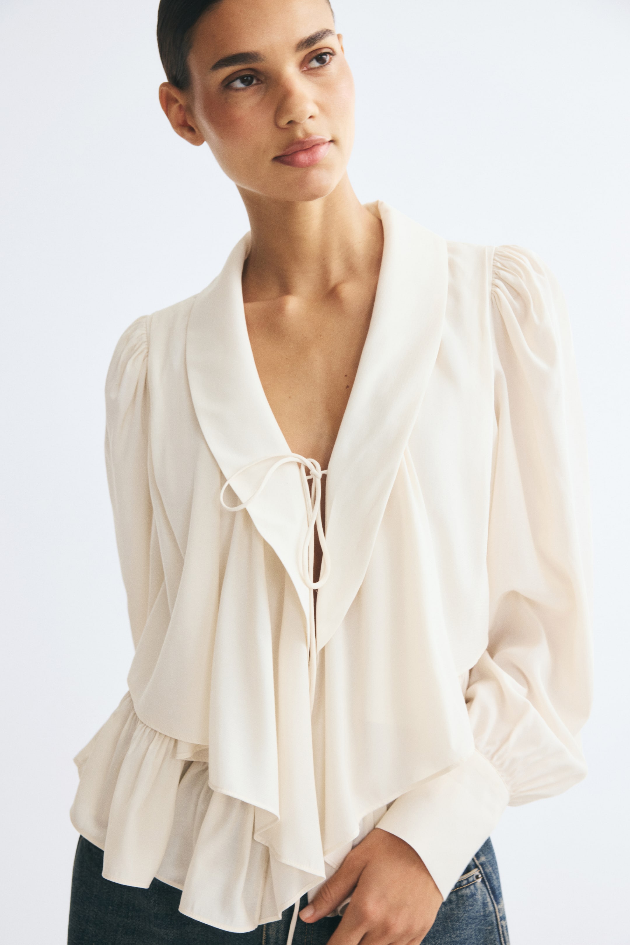 View larger image: The model is wearing an off-white blouse featuring a deep shawl collar with front spaghetti ties. The blouse is styled with blue jeans and has an open front with draped flounced tiers, long puff sleeves with wide cuffs, and a flared peplum hem.