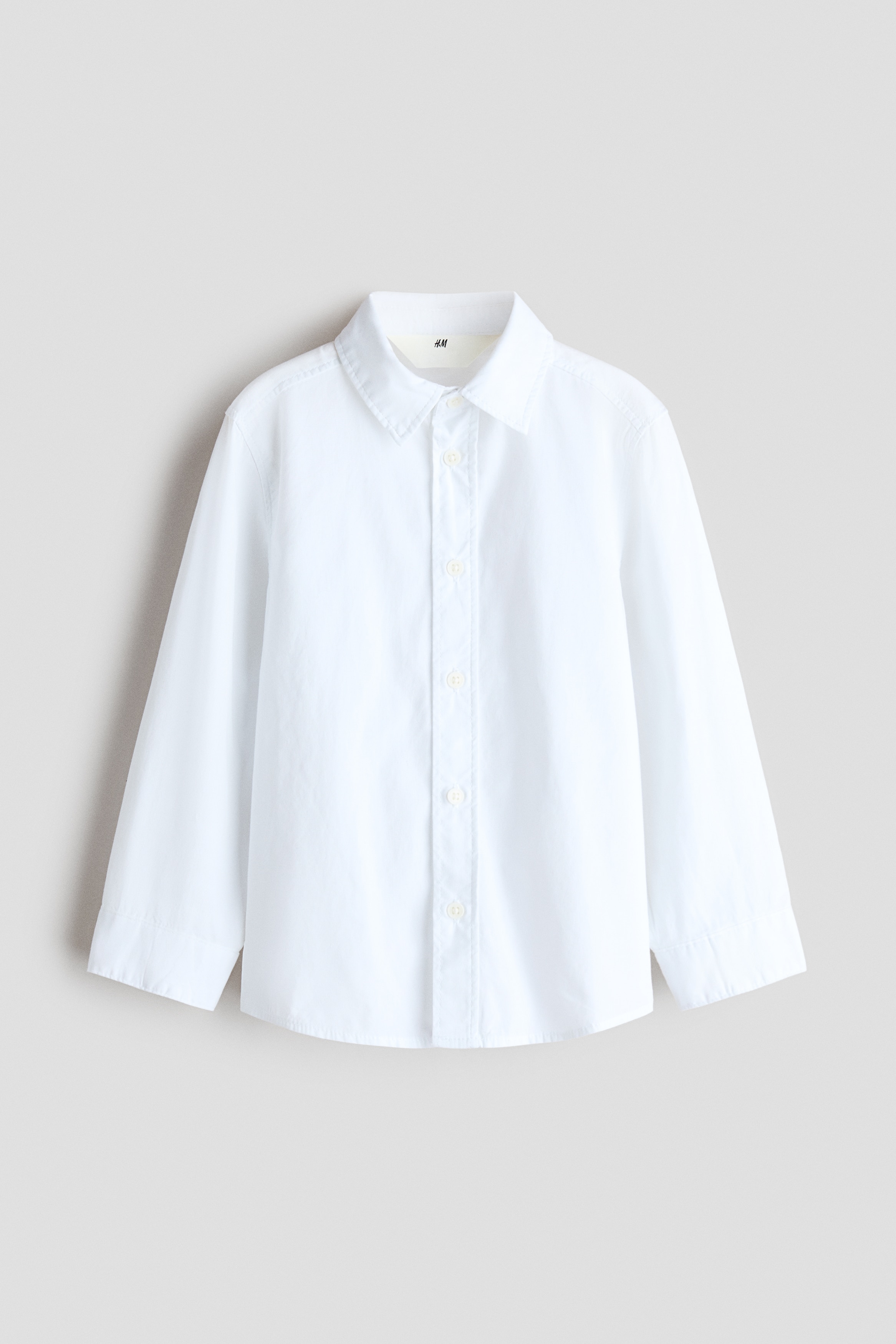 Una camicia bianca a maniche lunghe presenta un classico colletto a punta e una chiusura frontale con bottoni, che mostra chiaramente il suo design dalla parte anteriore. Si presenta con un taglio rilassato e maniche lunghe che terminano con polsini abbottonati.