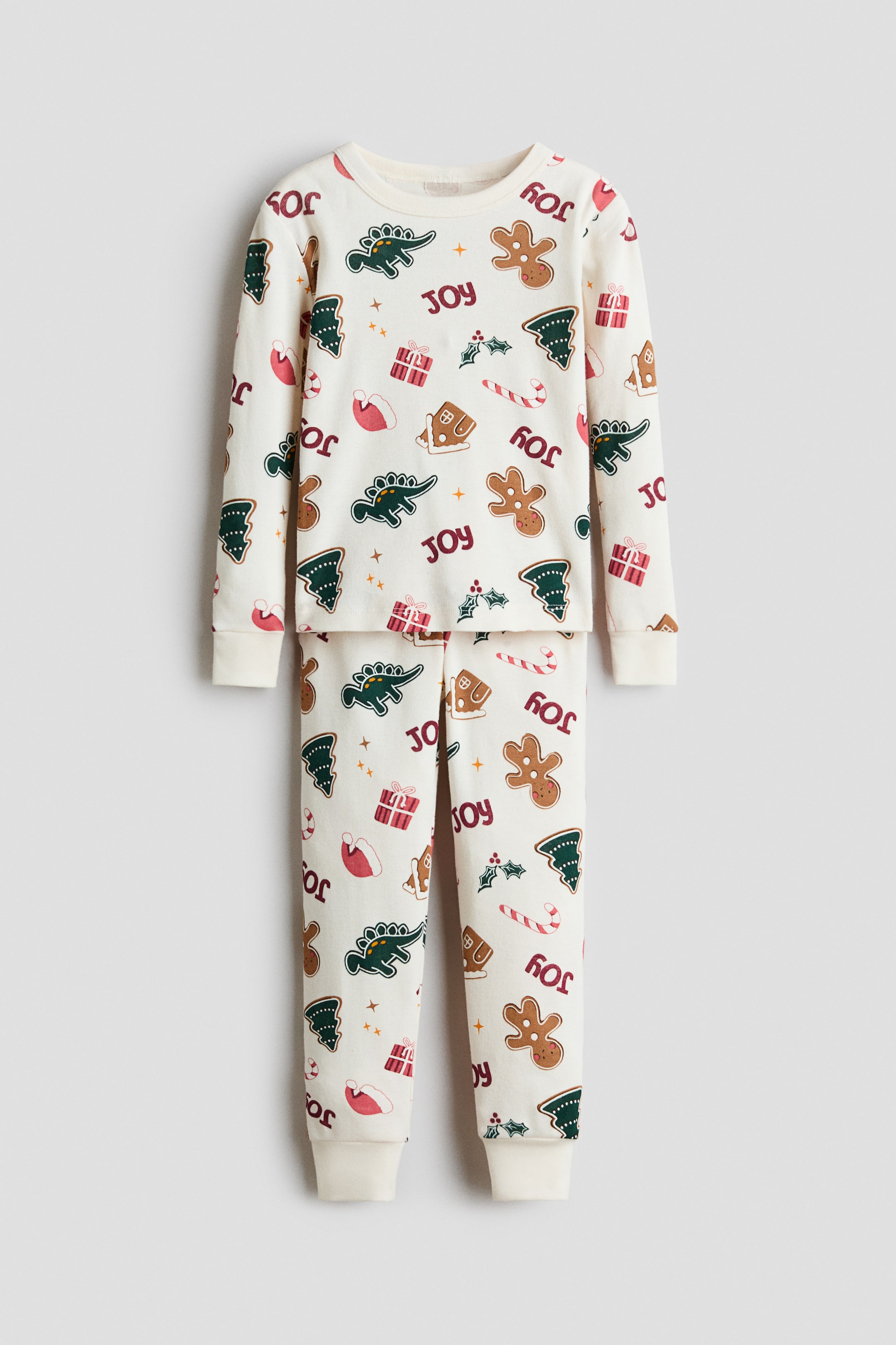 View larger image: A front-facing, two-piece pajama set in off-white is showcasing a festive holiday print with dinosaurs, gingerbread men, and the word "JOY." The long-sleeve shirt features a narrow-trimmed round neck and ribbed cuffs, while the matching pants display ribbed ankle hems.
