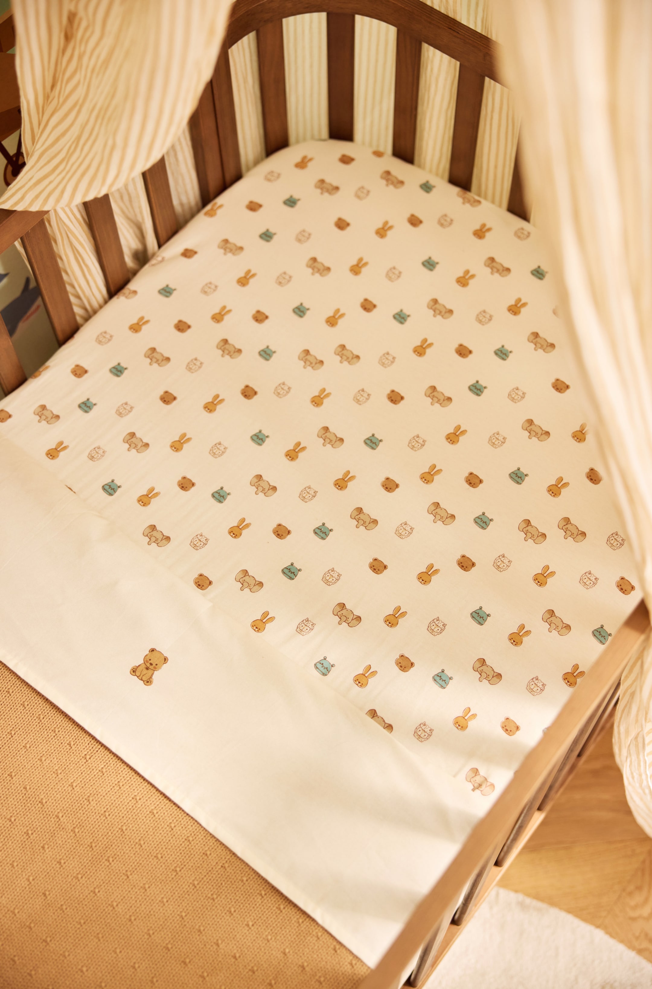 Grotere afbeelding bekijken: Een babybeddengoedset in crèmekleur bedekt een matrasje voor een kinderbedje en heeft een herhalend patroon van kleine, geïllustreerde dieren, waaronder beren, konijnen en olifanten, in zachte tinten. Het bijpassende vlakke laken is omgevouwen en toont een effen crèmekleurige omslag met een enkel geborduurd teddybeertje.