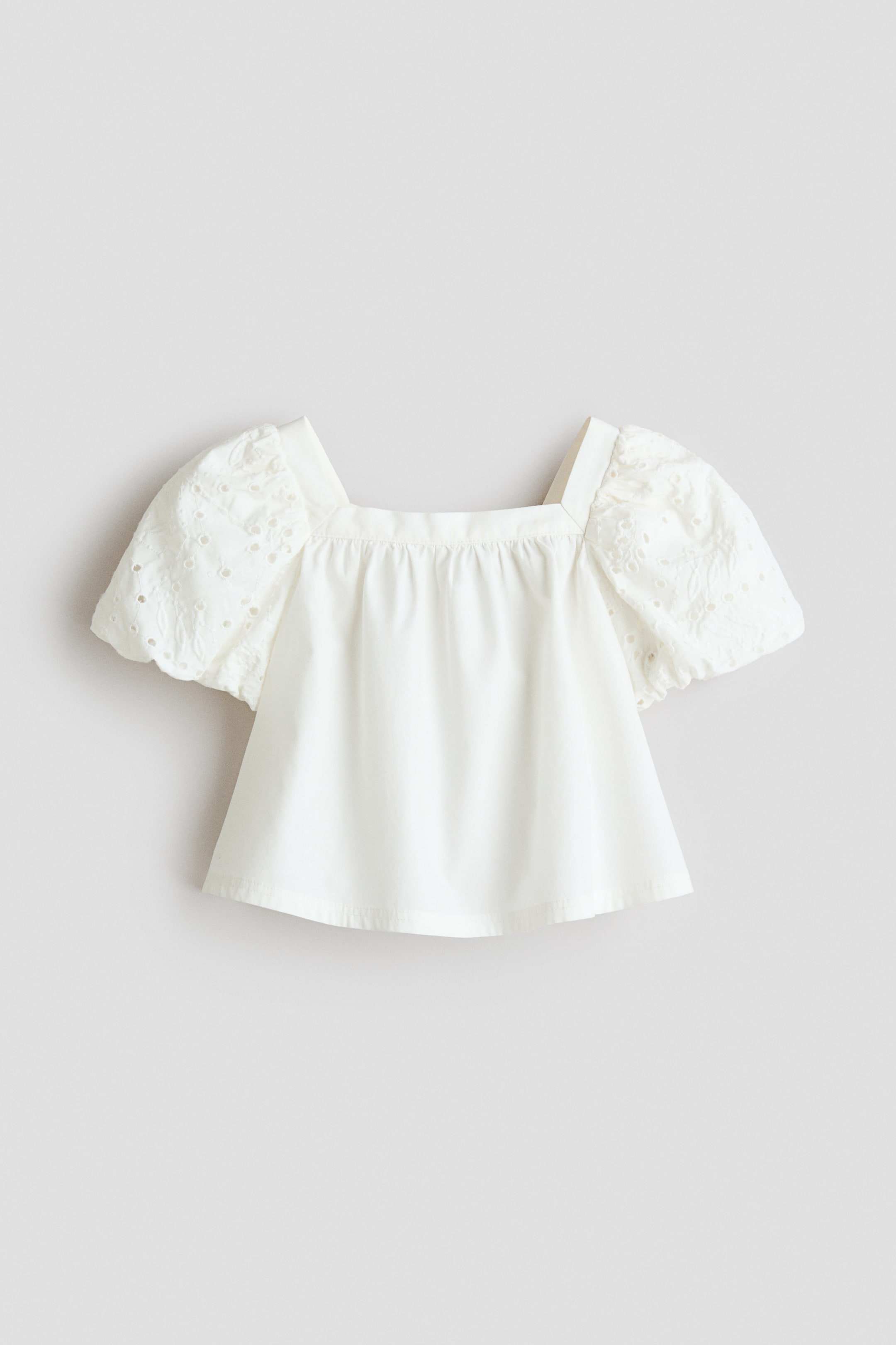 View larger image: A white top is displayed flat, facing forward, featuring a square neckline and short puff sleeves. The sleeves are adorned with eyelet embroidery, and the bodice is gathered at the neckline.
