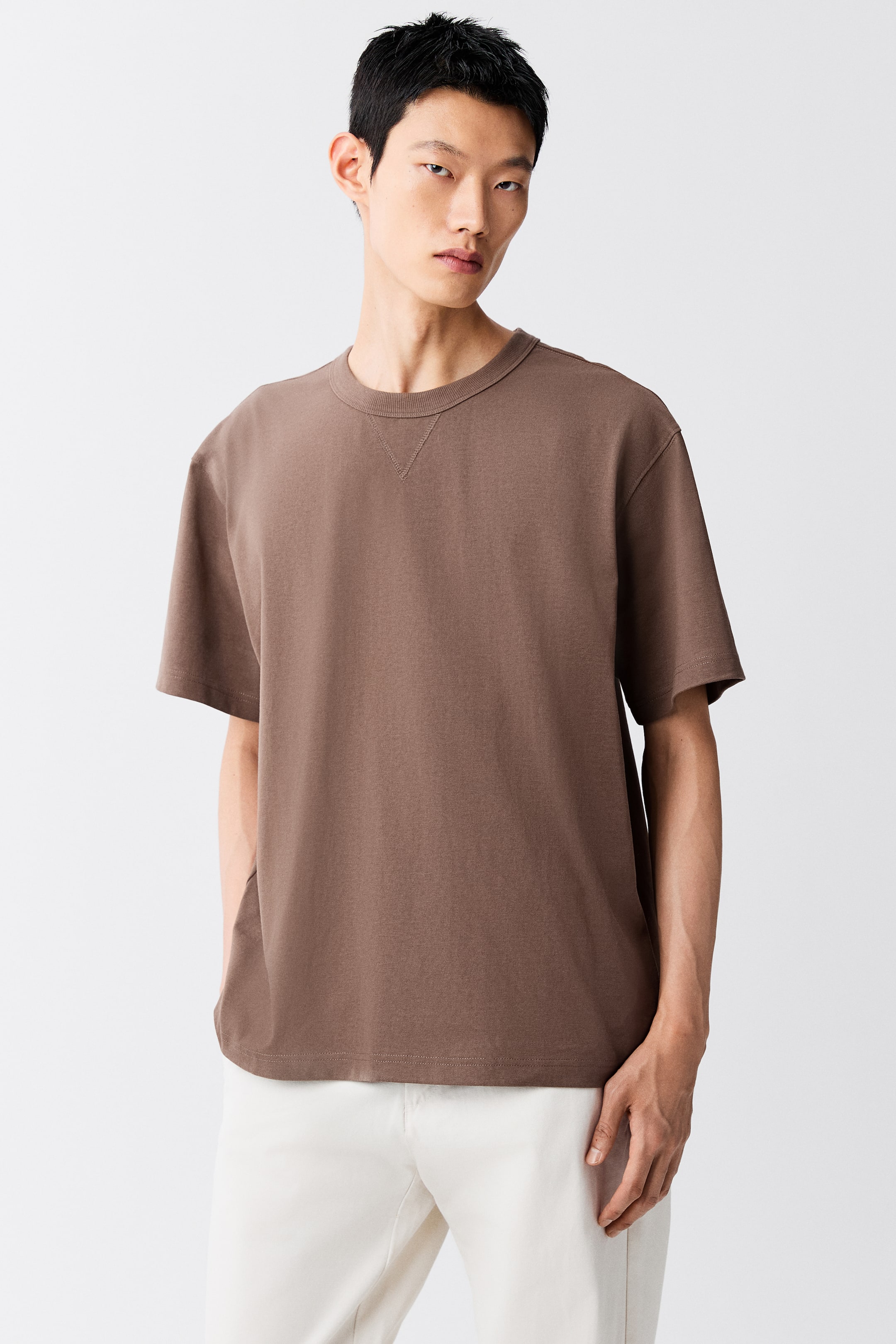 拡大画像を表示: モデルが着用しているのは、ブラウンのヘビーウェイトコットンジャージーTシャツ。ゆったりとしたフィット感、ドロップショルダー、三角形のステッチディテールをあしらったリブ仕様のクルーネックが特徴です。Tシャツはオフホワイトのトラウザーズとコーディネートされています。
