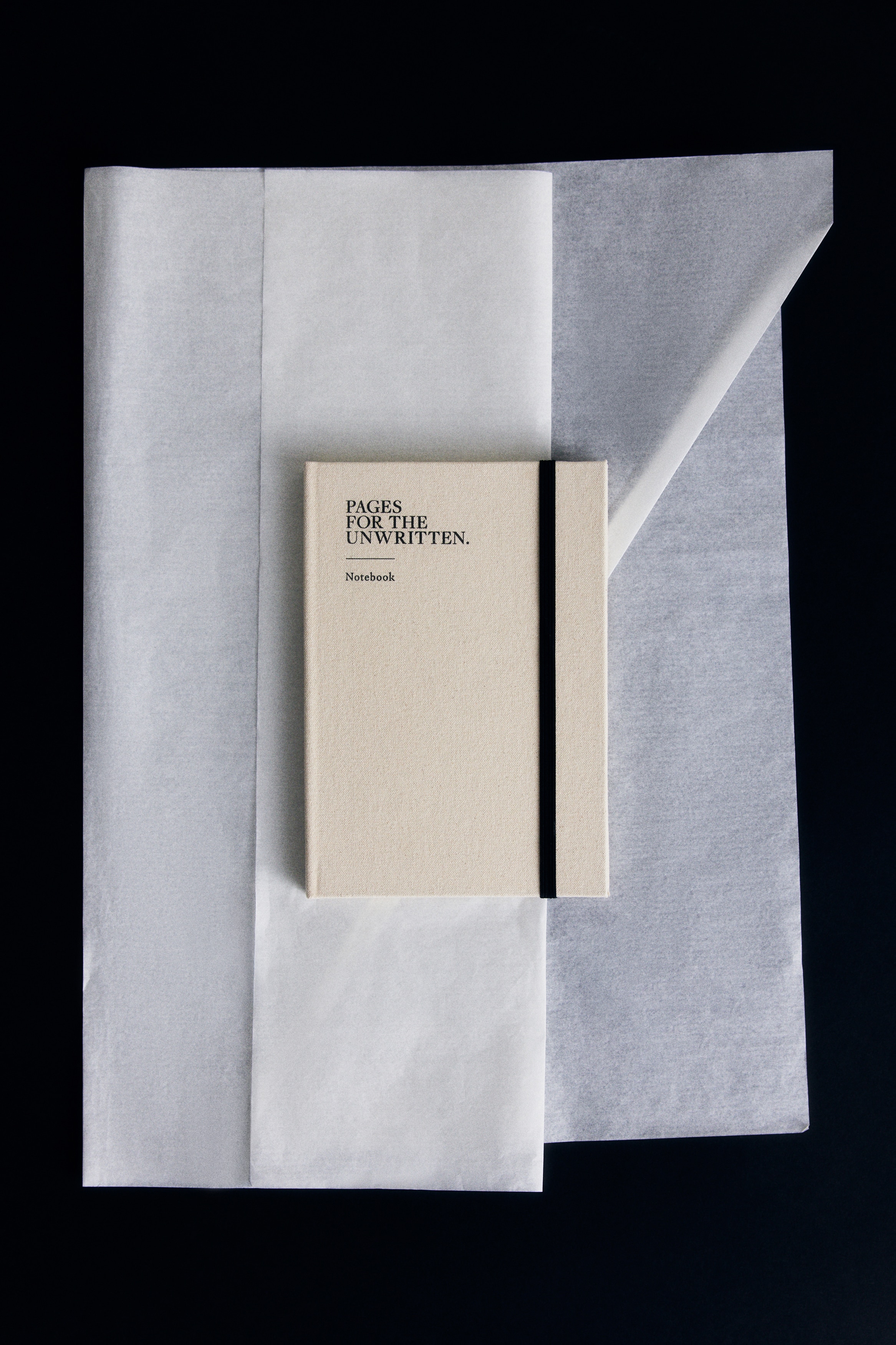 En krämfärgad, tyginbunden anteckningsbok visas framifrån, med den svarttryckta texten "PAGES FOR THE UNWRITTEN." och "Notebook" uppe till vänster på omslaget. Ett vertikalt svart elastiskt band löper längs högra sidan av omslaget för att hålla det stängt.
