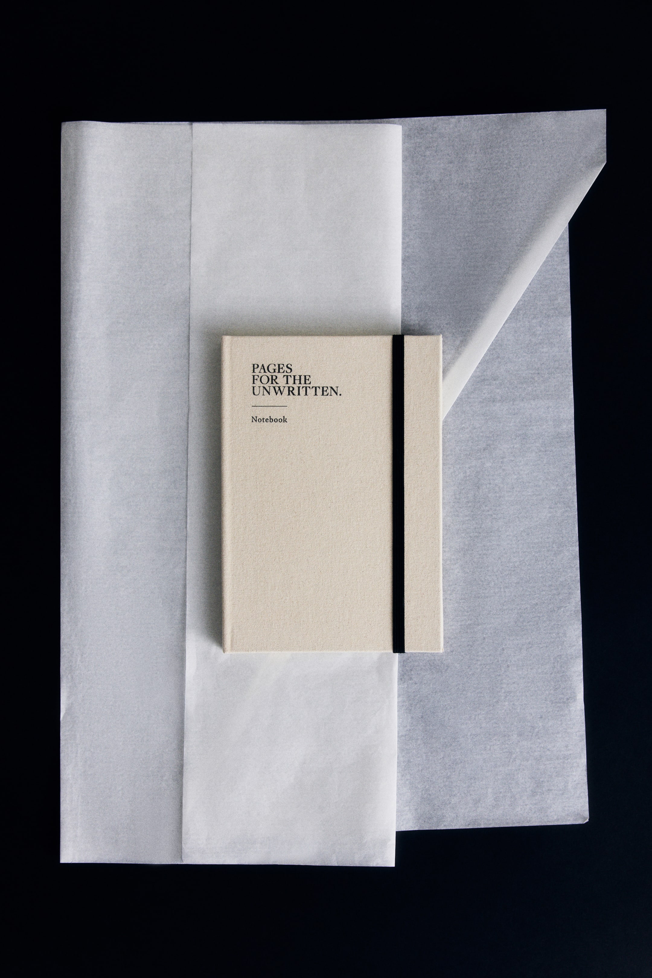 Se større billede: En beige notesbog med stofstruktur ligger fladt med forsiden opad på lag af hvidt silkepapir og har teksten "PAGES FOR THE UNWRITTEN." og "Notebook" trykt i sort øverst til venstre på omslaget. Et sort elastikbånd løber lodret nær højre kant og holder notesbogen lukket.