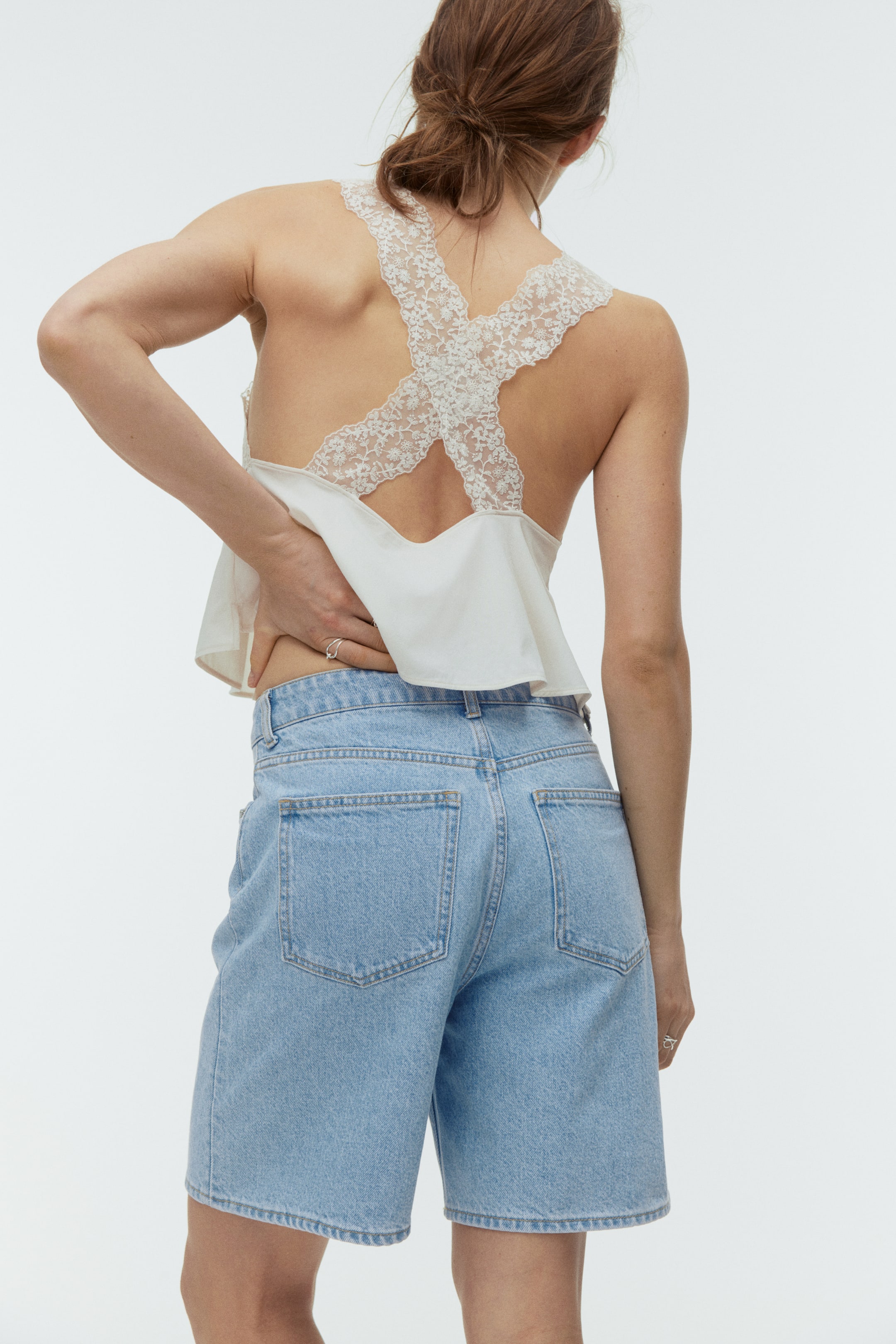 View larger image: An off-white camisole top features a flowy back and criss-cross lace straps, paired with light blue denim high-waisted shorts that fall to the knee and show back patch pockets, viewed from the back.