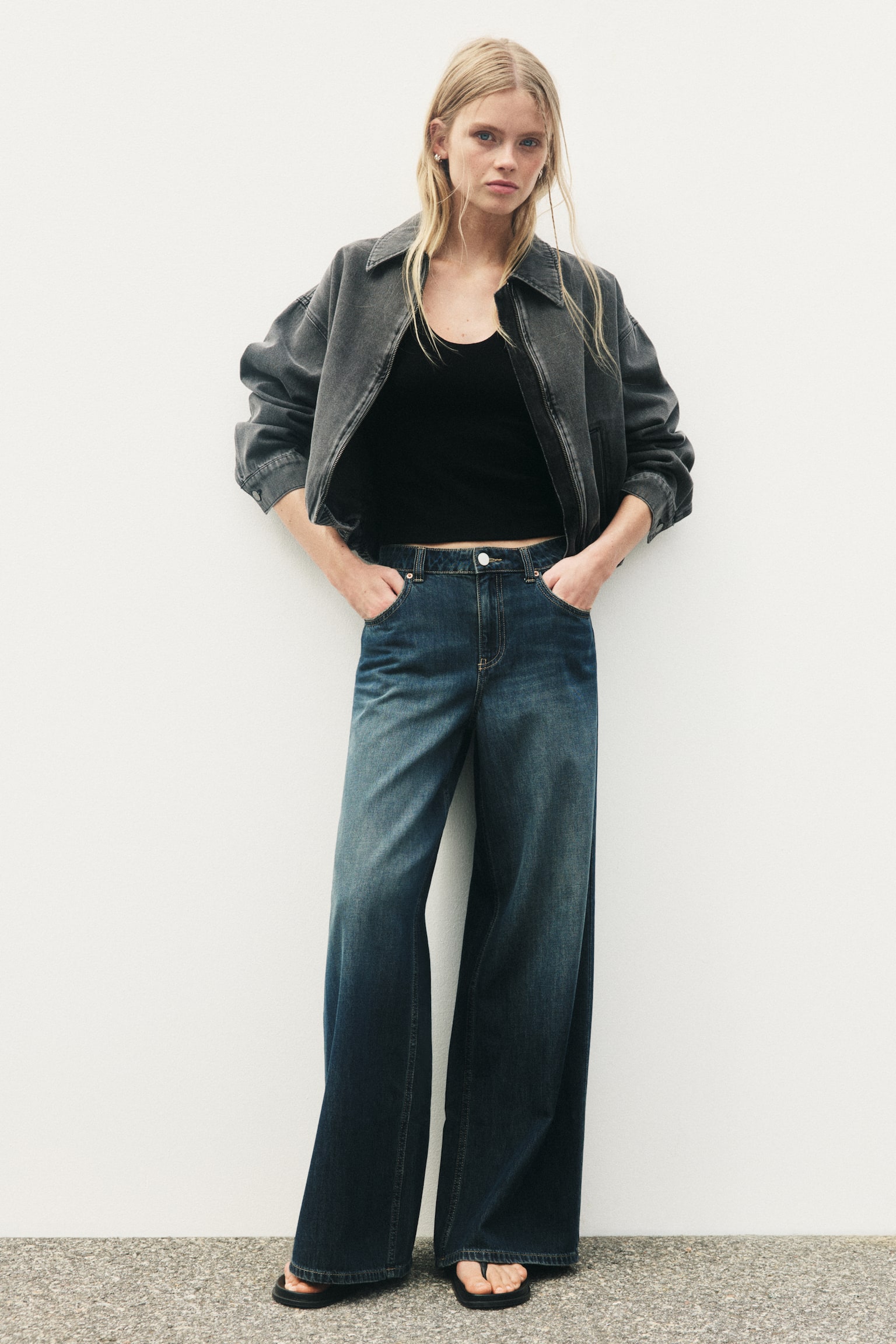 Wide-leg, regular-waisted jeans in a faded dark blue wash are being worn by the model, who is standing with her hands in the front pockets. The jeans feature a five-pocket design and a wide cut from the thighs to the hems, and are styled with a black crop top and an open grey denim jacket. - 1