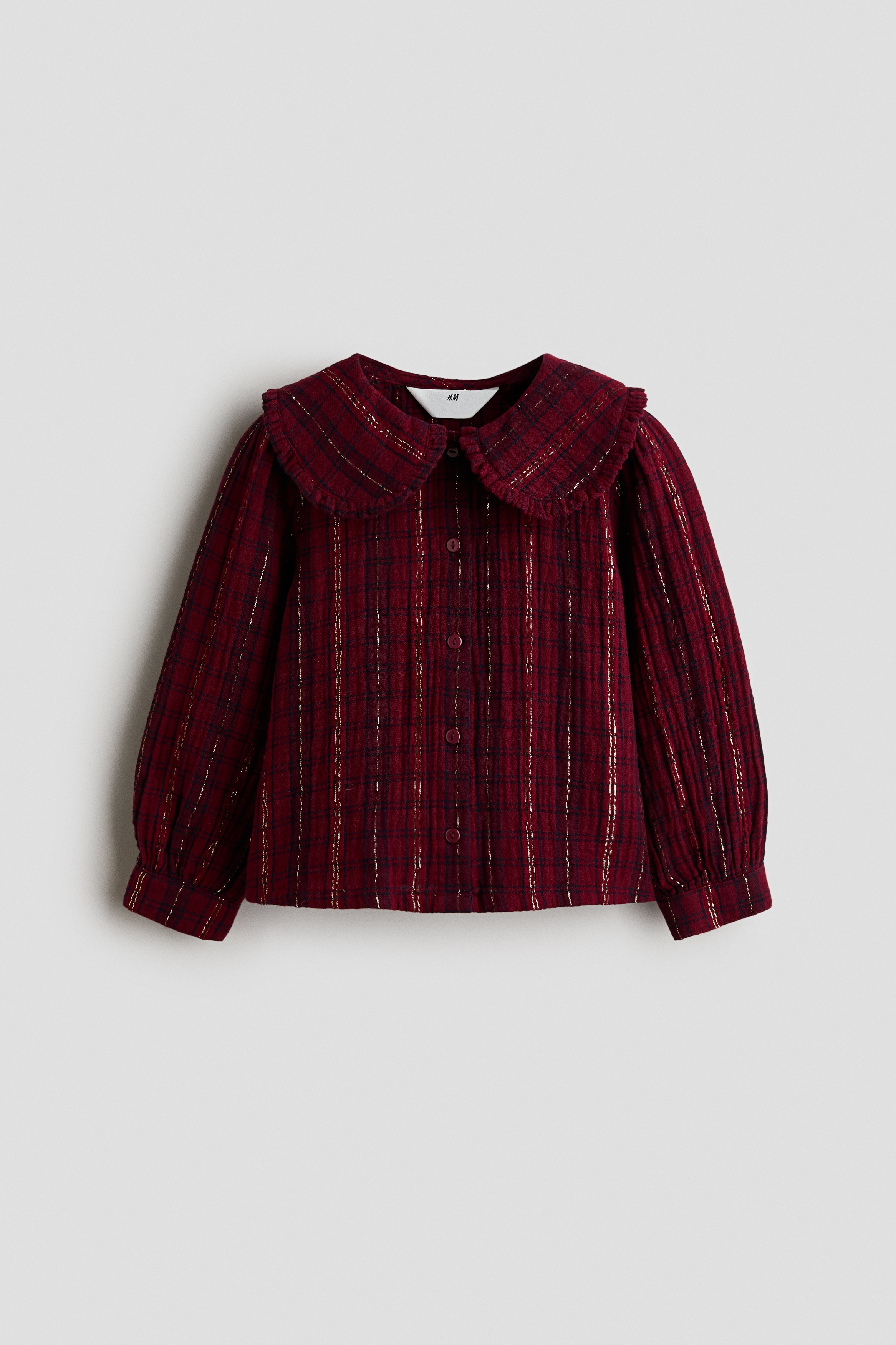 Una vista frontale mostra una blusa rosso scuro in tessuto jacquard di cotone, caratterizzata da un motivo tartan con fili scintillanti glitterati, un colletto Peter Pan rifinito con volant e una chiusura frontale con bottoni. La blusa è abbinata a una gonna tartan coordinata, mettendo in risalto il suo orlo dritto e la vestibilità morbida.
