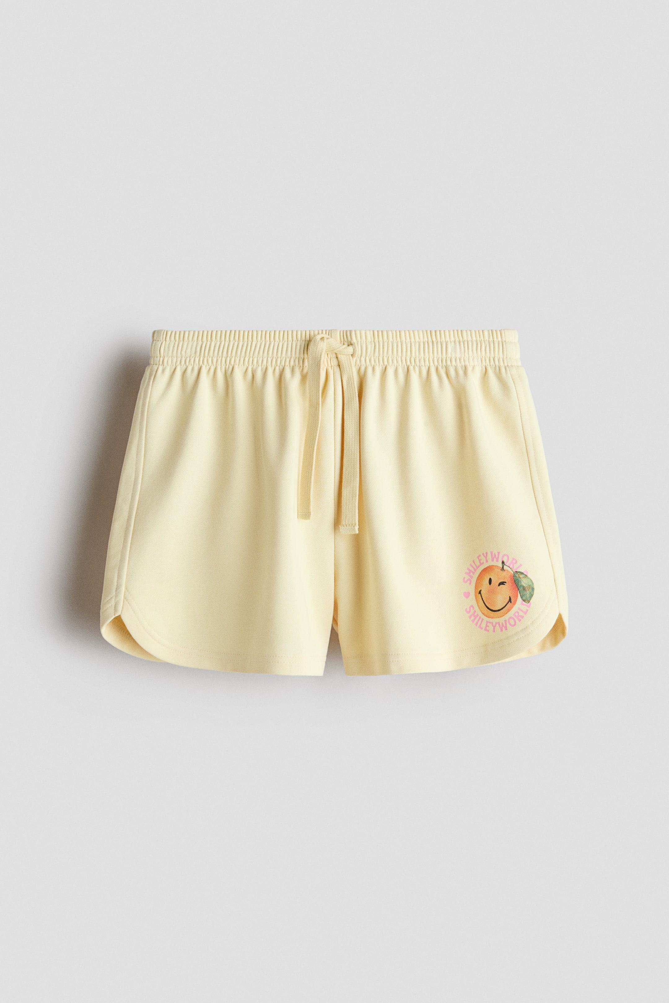 View larger image: Front-facing, light yellow relaxed-fit shorts in sweatshirt fabric featuring an elasticated drawstring waist with visible ties, a circular winking orange smiley face print on the lower right leg, and gently curving hems.