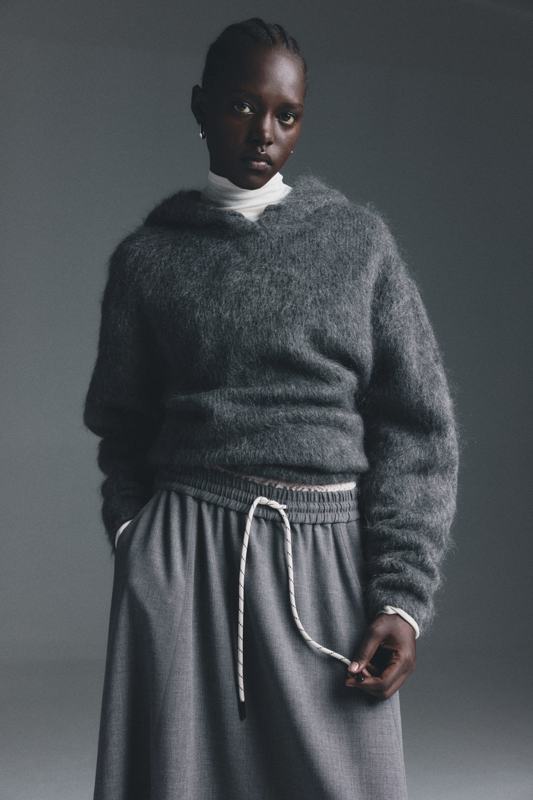 View larger image: A dark gray fuzzy knit hooded sweater is being worn over a white turtleneck, presenting a relaxed fit. It is being paired with gray wide-leg pants, which are designed with an elasticated waistband and a visible white and black speckled drawstring that a hand is holding.