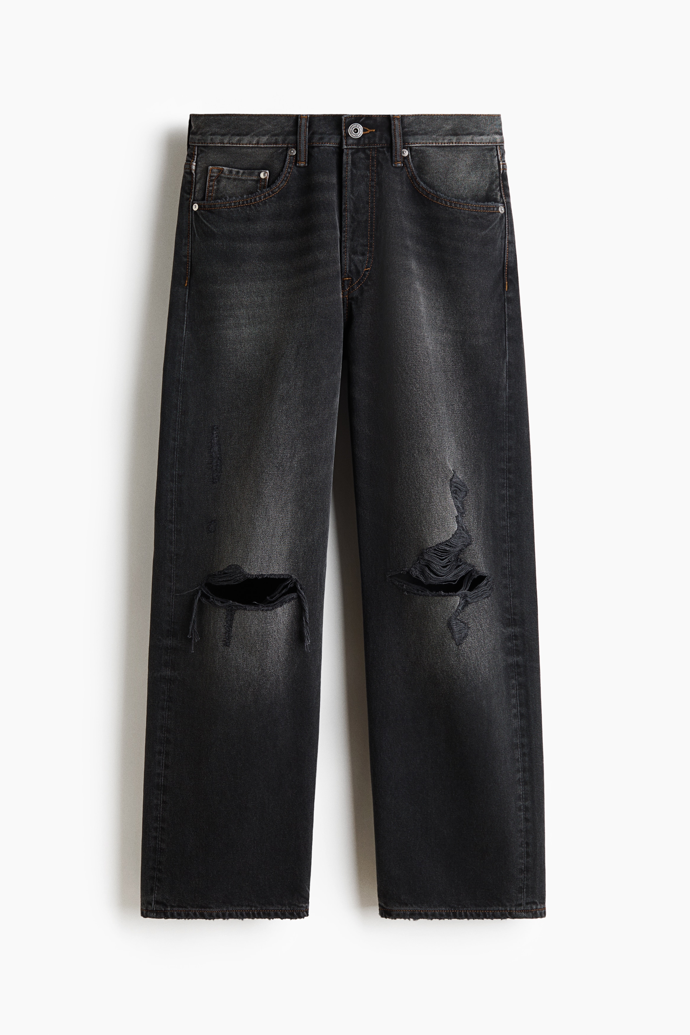Dark grey, loose-fit denim jeans featuring a straight-leg cut and distressed, ripped details at the knees are worn by the model. The jeans are paired with a tucked-in black long-sleeve Henley shirt and black shoes, with the model resting one hand in a front pocket.
