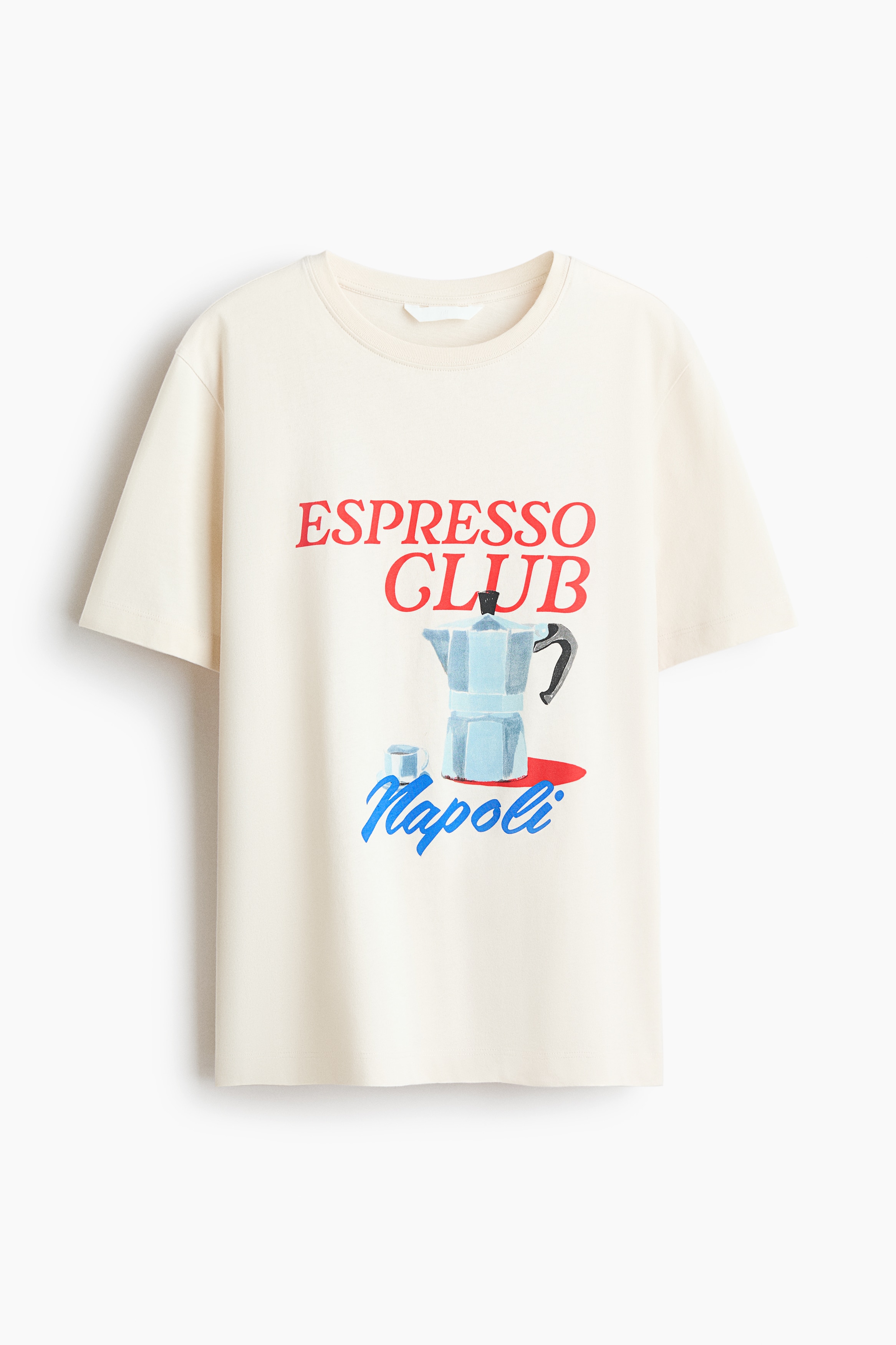 A light beige T-shirt featuring a round neckline, gently dropped shoulders, and a front graphic print of a moka pot with the text "ESPRESSO CLUB Napoli" is being worn by the model. The tee is layered over a white T-shirt and paired with light-coloured jeans.
