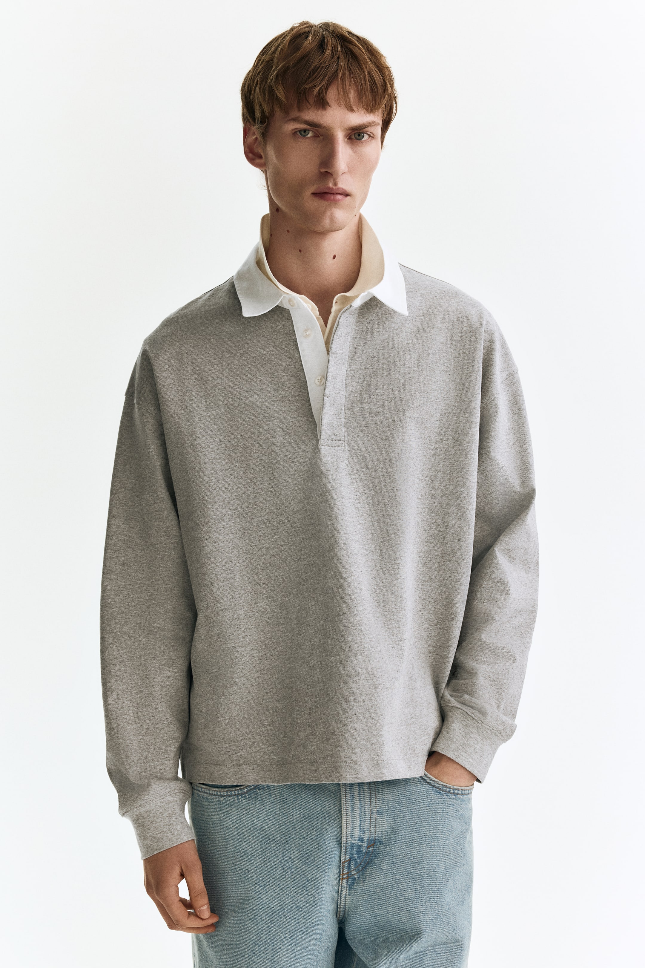 הצגת תמונה גדולה יותר: חולצת פולו אפורה עם שרוולים ארוכים כוללת צווארון פולו לבן המונח על גבי צווארון חולצה בצבע בהיר, ושלושה כפתורים לבנים. היא מציגה גזרה רפויה מקדימה, עם חפתים מחורצים.