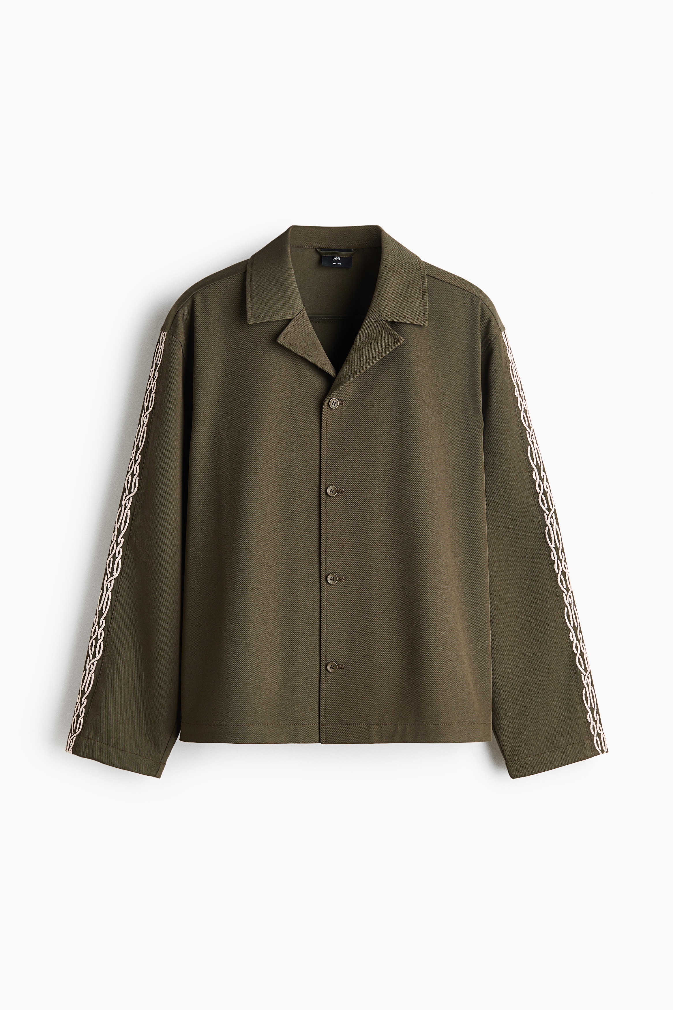 The model is wearing an olive green overshirt featuring a resort collar, a buttoned front, and a white embroidered chain motif along the sleeve. The shirt is styled with matching trousers as the model stands in a three-quarter profile.