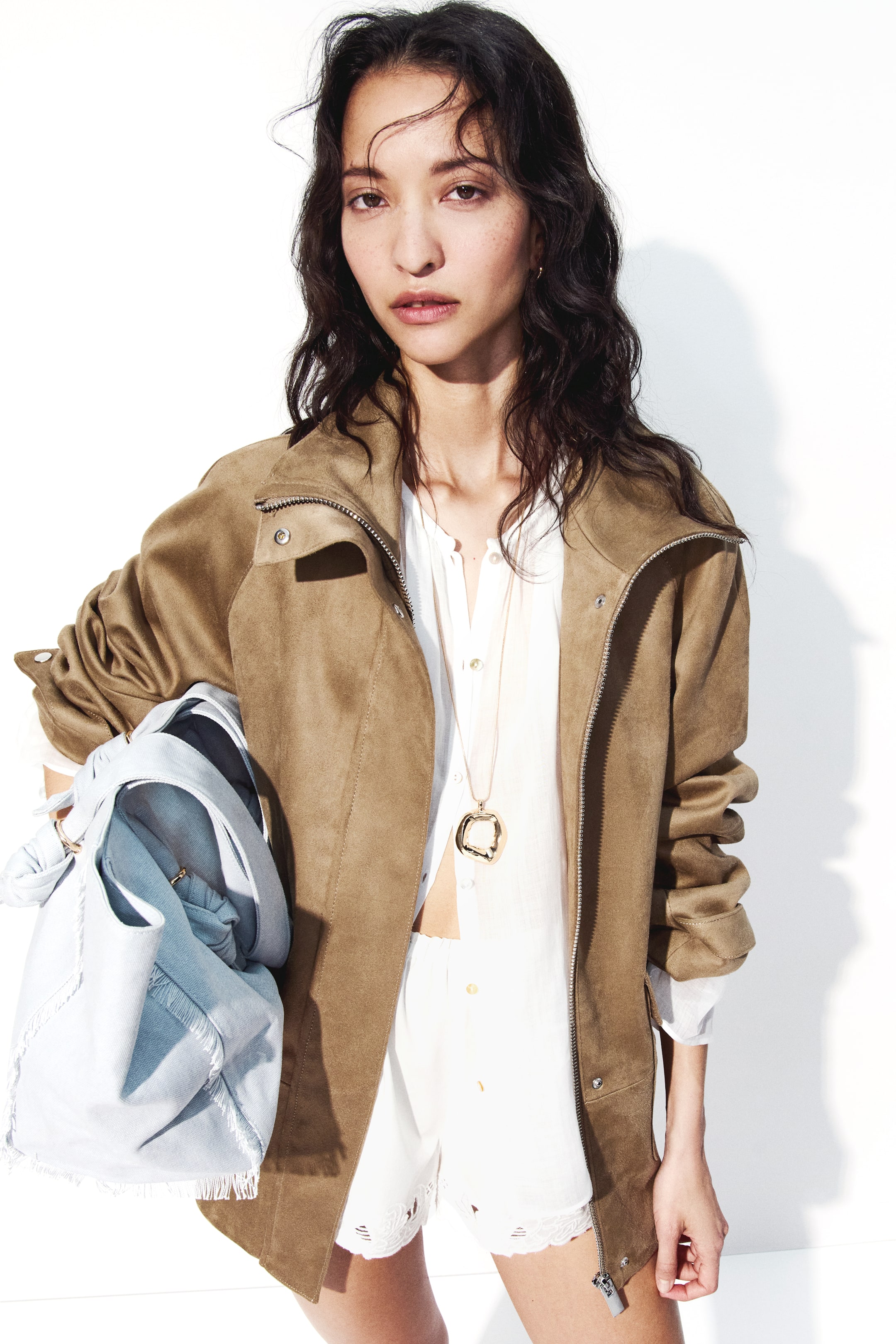 Agrandir l'image: Une veste oversize avec une finition douce et duveteuse et un col montant est portée ouverte par le mannequin, révélant une fermeture éclair et un rabat coupe-vent sur le devant ainsi que des manches raglan. Le mannequin tient un sac bleu clair sous son bras gauche, qui repose contre la veste.