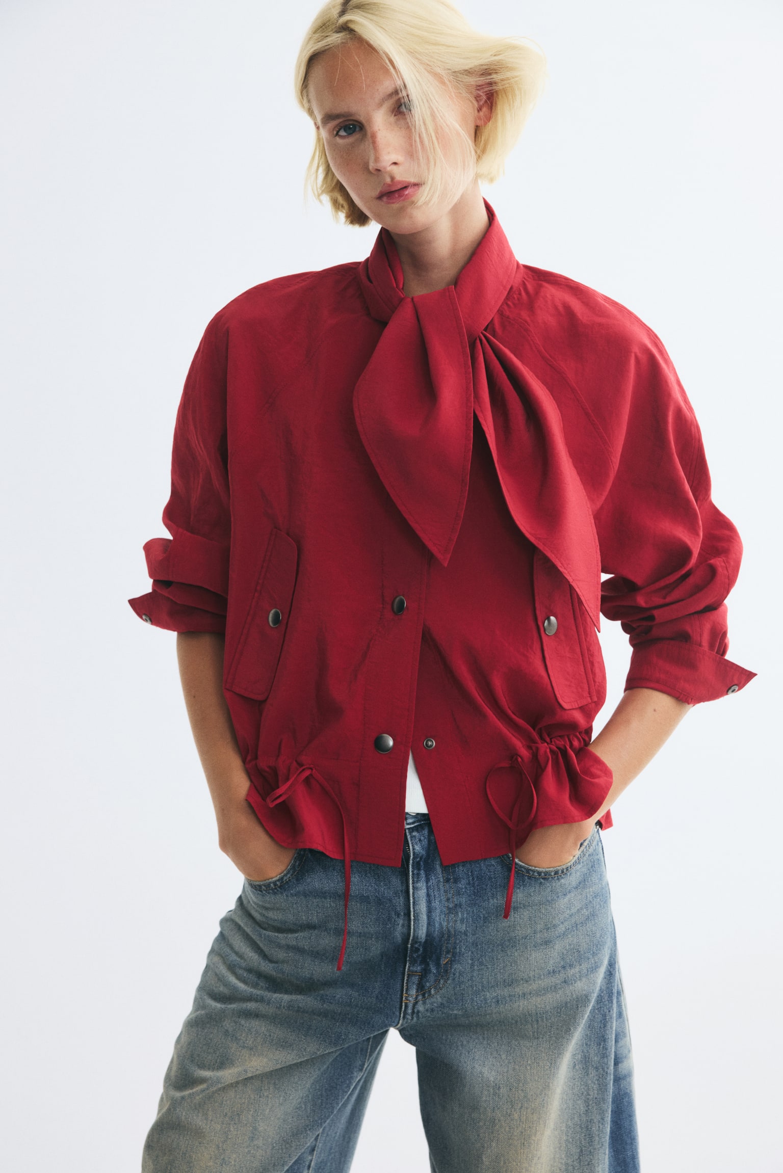 A red jacket is shown from the front, featuring a high collar with an integrated, knotted tie-neck scarf and a central snap-button closure. The jacket has long sleeves, slanted flap pockets with snap closures, and a gathered drawstring hem with thin ties. - 1