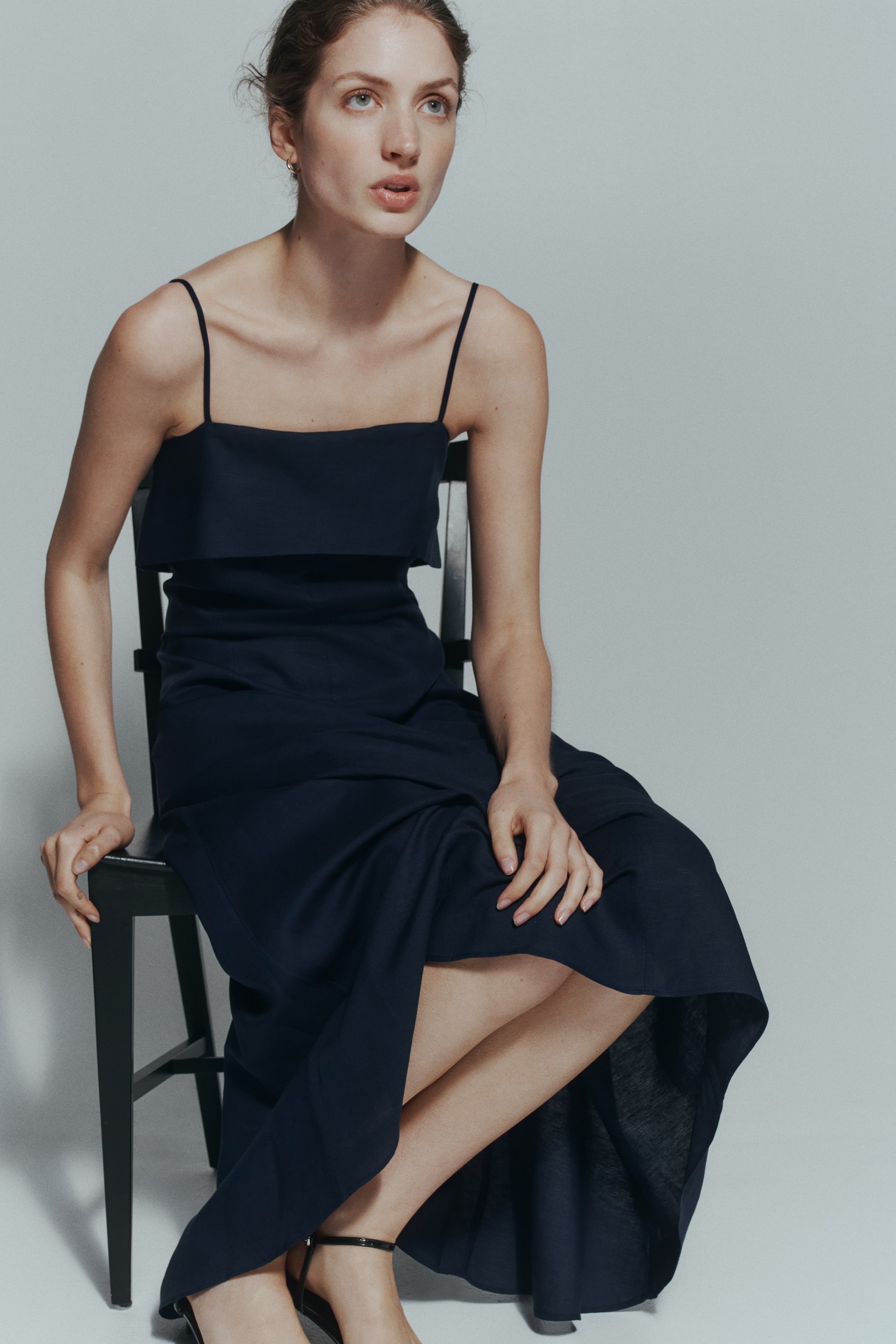 View larger image: The model is wearing a dark blue calf-length dress in an airy weave, featuring spaghetti shoulder straps and a wide, straight foldover top edge. Its fitted bodice transitions into a gently flared skirt that drapes around her legs as she sits on a chair, paired with black strappy sandals.