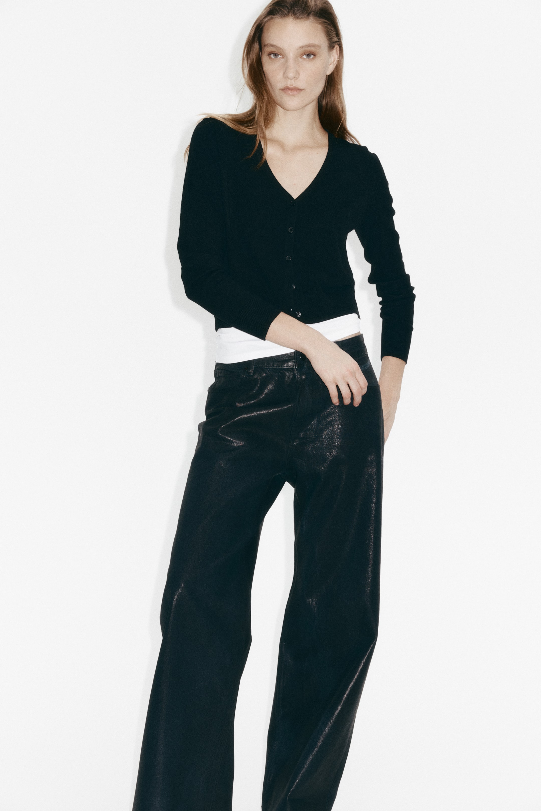 View larger image: A black V-neck cardigan is worn, featuring long sleeves, a button-front closure and a cropped length, layered over a white top. Black wide-leg trousers are also worn, showcasing a crinkled, slightly shiny texture and a relaxed fit.