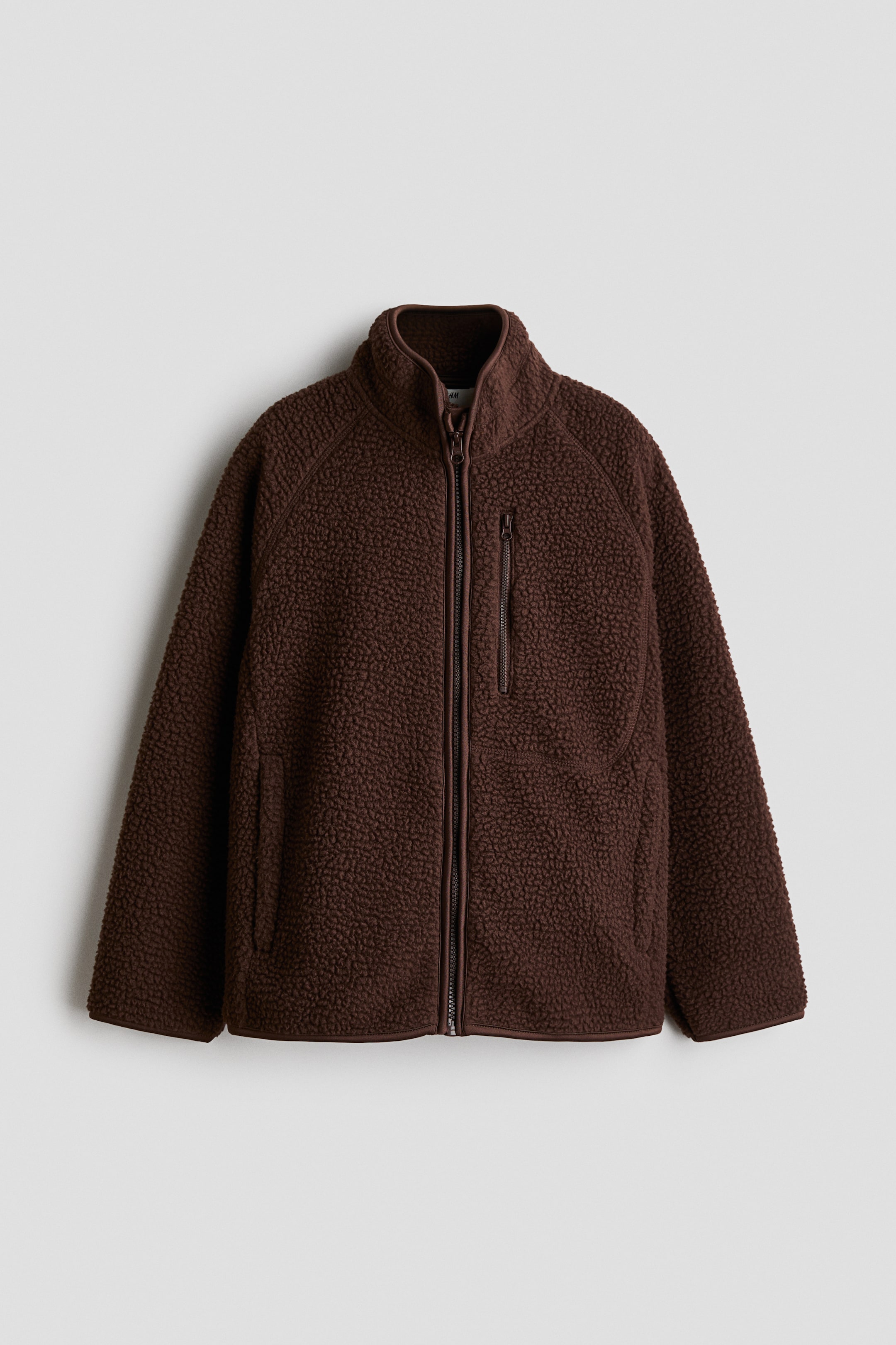 Voir une image plus grande: Une veste en polaire peluche marron est dotée d'un col montant et d'une fermeture éclair frontale sur toute la longueur. Elle présente également une poche poitrine zippée verticale du côté gauche et deux poches latérales, avec des coutures raglan définissant la structure des épaules.