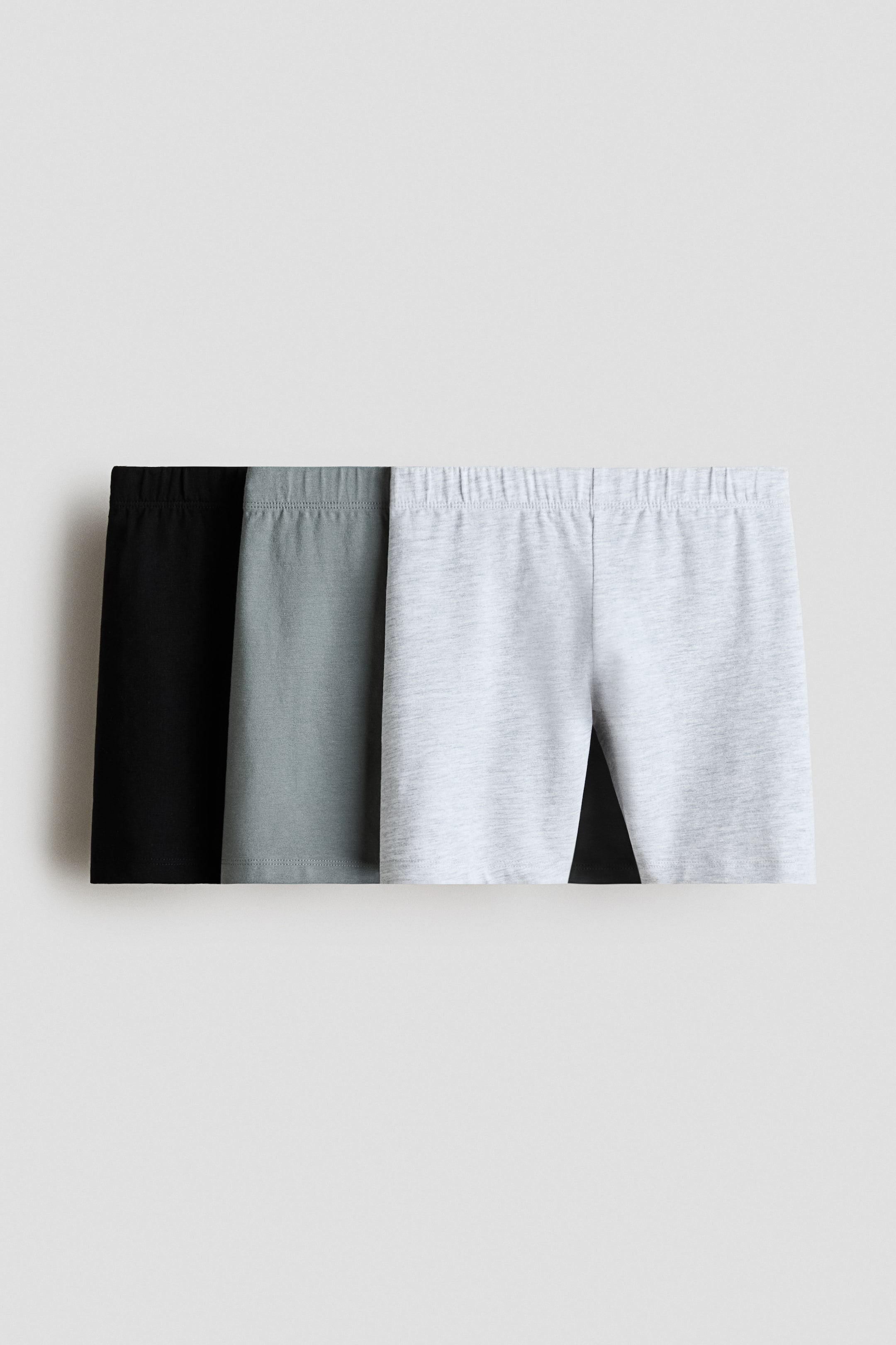 View larger image: Three biker shorts display their front elastic waistbands and plain knit fabric in black, dark gray, and light gray, layered from left to right.