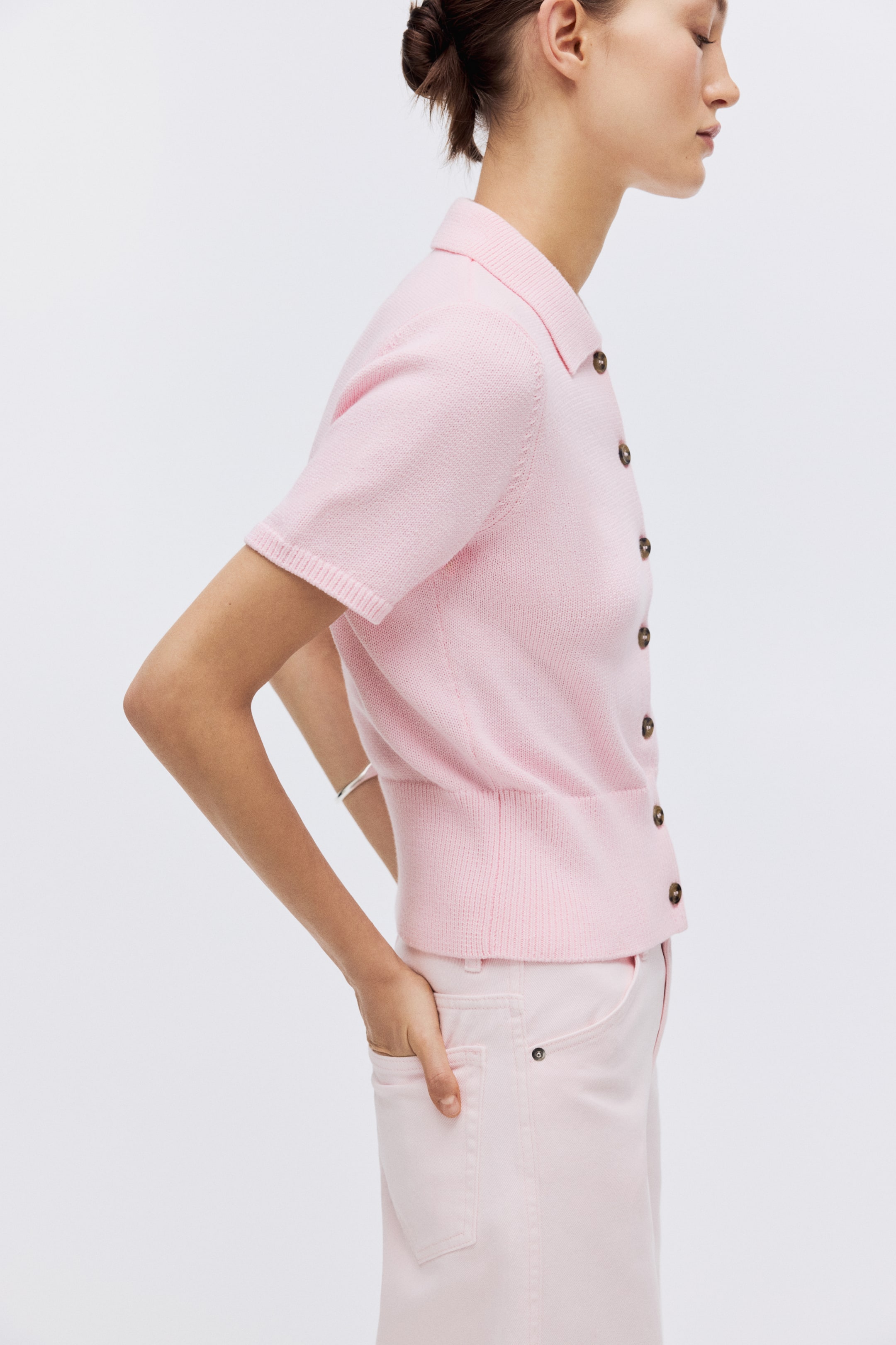 View larger image: A light pink knitted short-sleeved polo cardigan is shown in profile. It features a classic polo collar, a full button-front fastening with dark buttons, and ribbed short sleeves and hem.