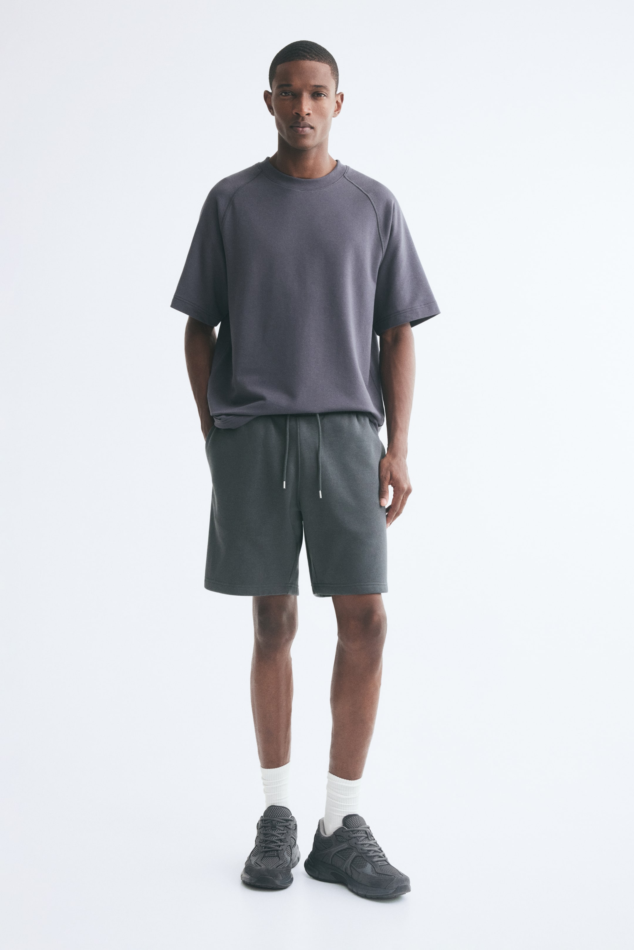 View larger image: Sweat shorts in a dark grey shade are being worn by the model, featuring a relaxed fit, an elasticised waistband with an adjustable drawstring, and side pockets. The model is standing with one hand resting in a pocket, styling the shorts with a dark grey T-shirt, white ribbed socks, and grey sneakers.