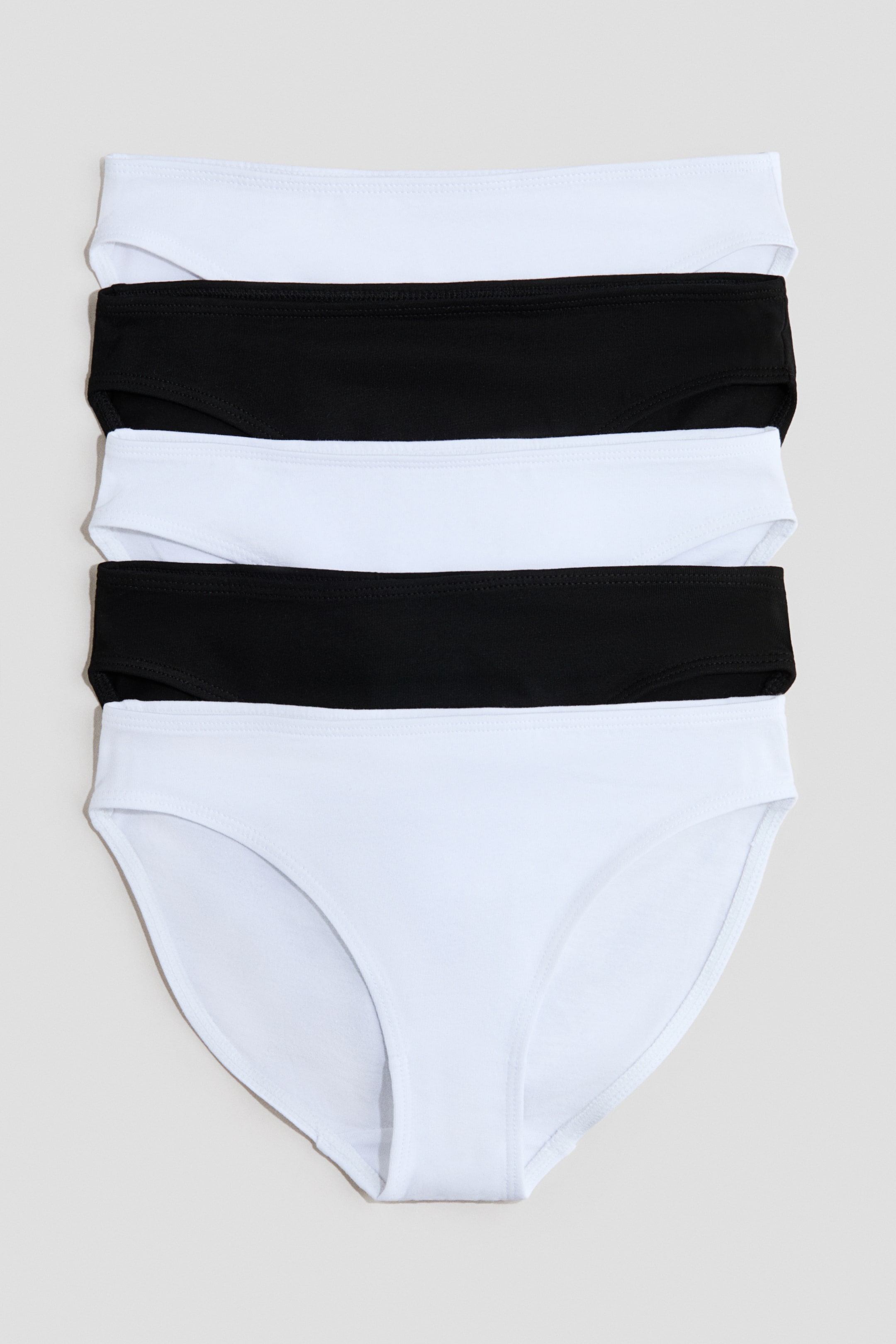 View larger image: Five women's bikini briefs are stacked neatly, alternating in colour between white and black, presented from a front view. Each brief features an elasticated waistband, with the bottom white brief also showcasing its high-cut leg openings.