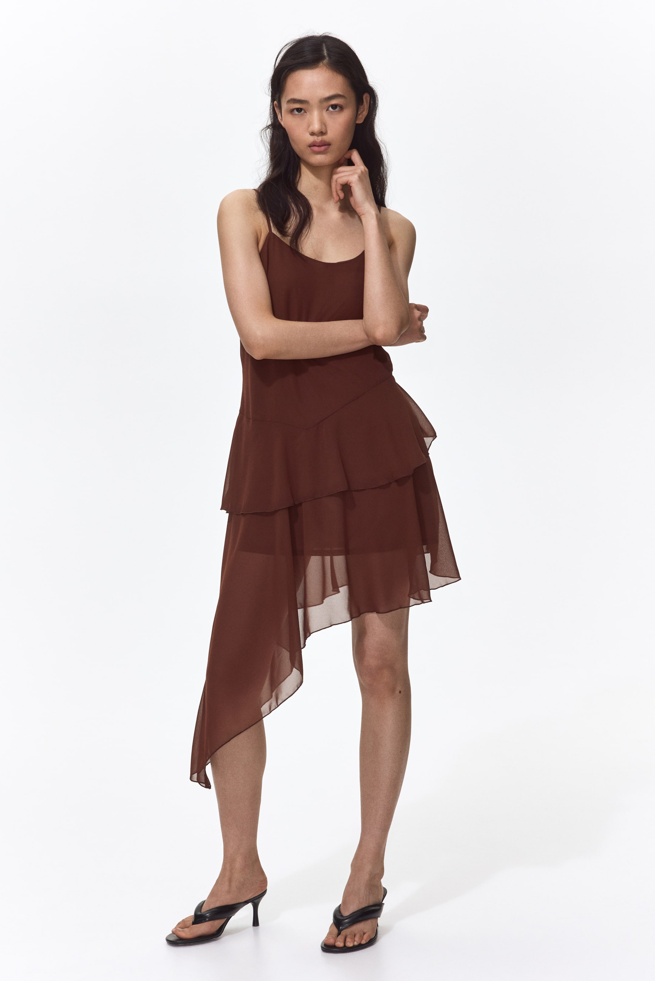 View larger image: A brown slip dress is featuring slender spaghetti straps and a scoop neckline. The tiered skirt showcases sheer ruffled layers and an asymmetrical hem, with one side extending notably lower than the other.