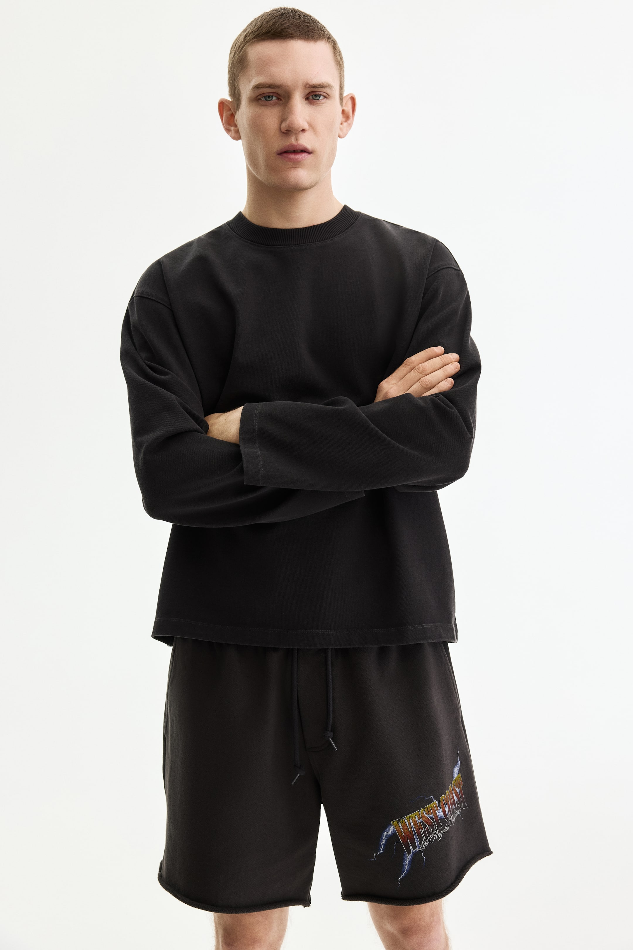 View larger image: A black crew-neck, long-sleeve top, front-facing the viewer, features a relaxed fit with wide, unribbed cuffs. It is paired with black shorts featuring a drawstring waist, raw-cut hems, and a colorful graphic print reading "WEST COAST" with lightning bolts on the left leg.