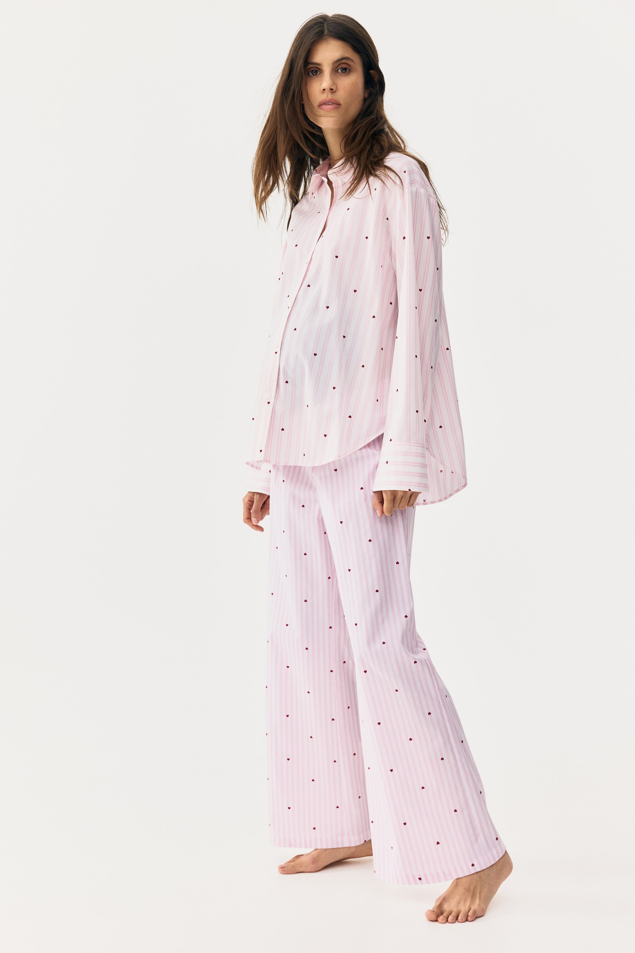 View larger image: A light pink pyjama set, featuring thin white vertical stripes and small red heart patterns, is showcasing a loose-fitting, long-sleeved button-up shirt with a collar and wide-leg trousers. The set is angled slightly to the right, displaying its front and side.