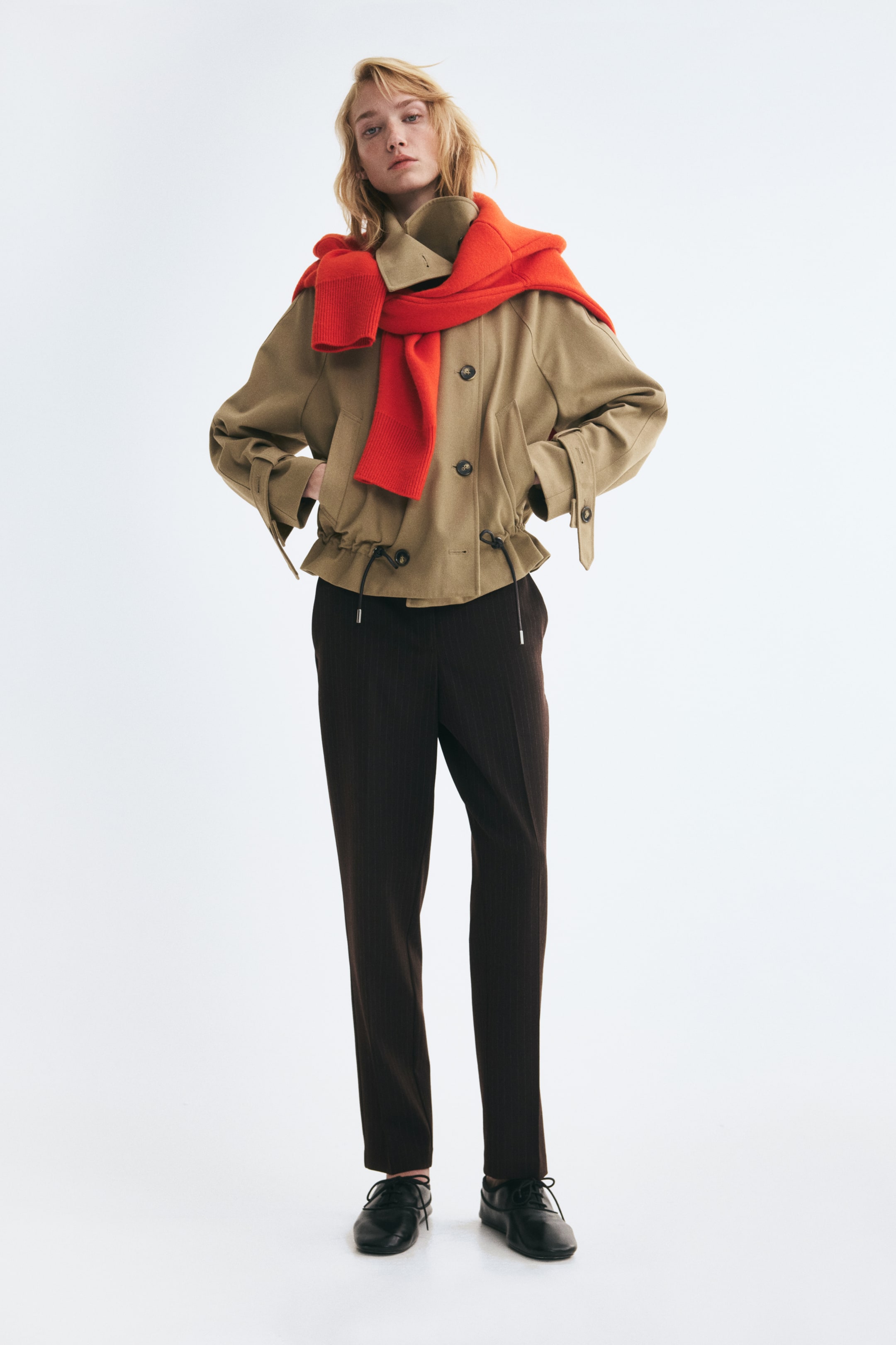 Agrandir l'image: Un pantalon cigarette marron foncé, longueur cheville, à jambe fuselée, est porté par le mannequin. Le pantalon est associé à une veste utilitaire beige et à des chaussures noires à lacets.