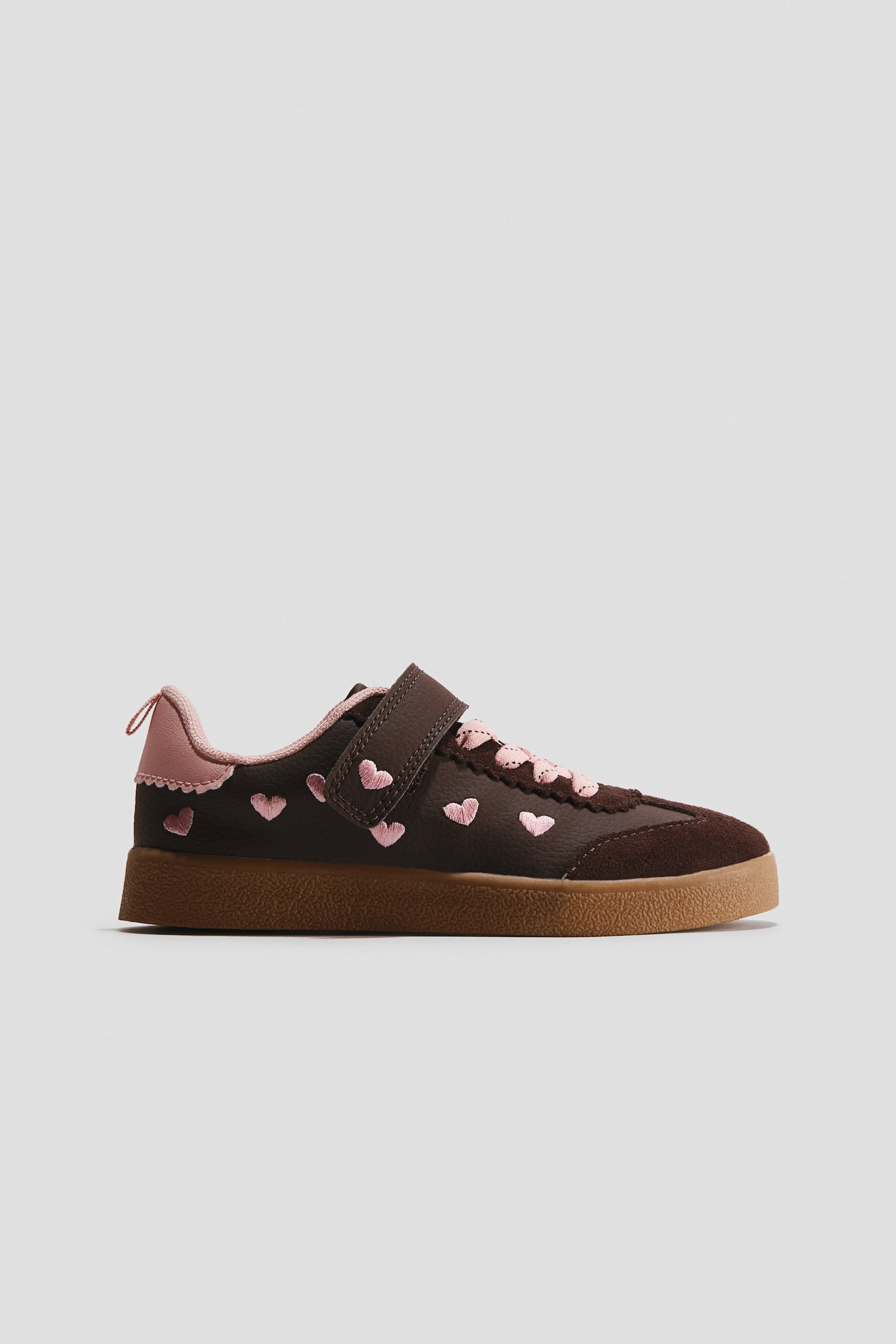 A retro-style sneaker is shown in profile, featuring a dark brown upper adorned with embroidered pink hearts and a hook-and-loop strap closure over pink laces. The shoe showcases a textured gum rubber sole, a pink heel tab, and scalloped pink detailing along the collar.