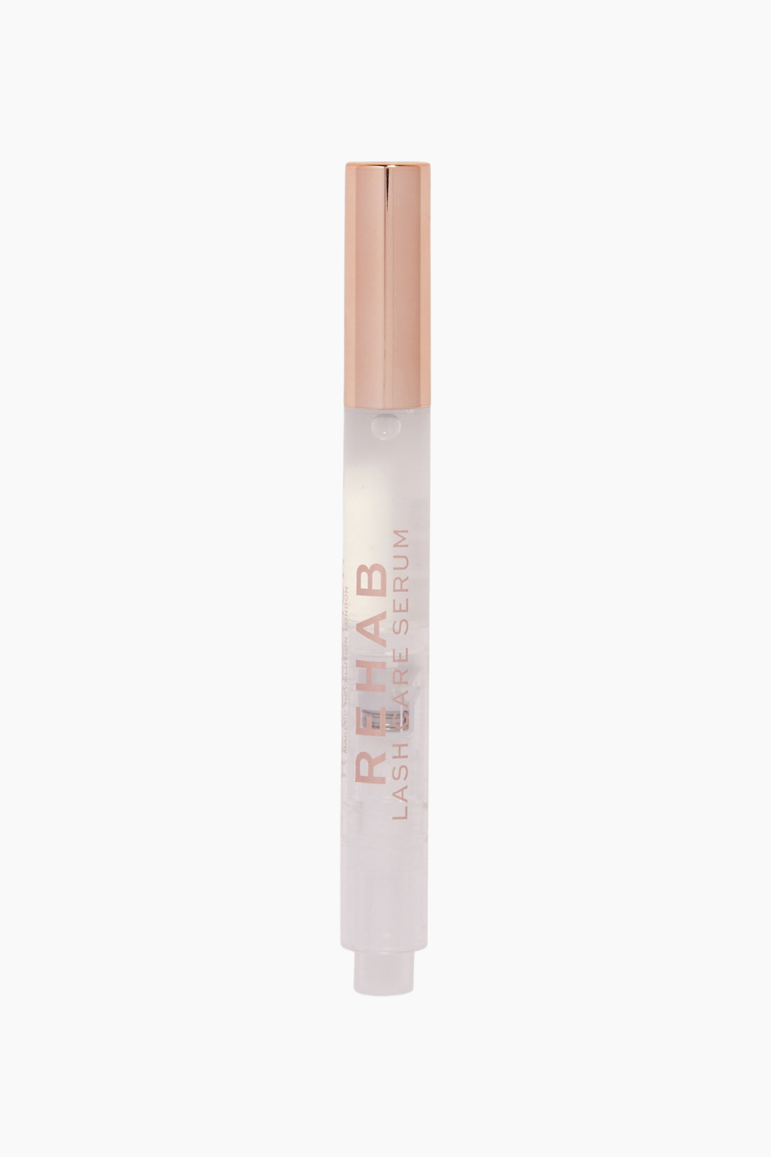 Revolution Rehab Lash Care Serum stÃ¥r oppreist, og har et klart sylindrisk rÃ¸r som viser en gjennomsiktig vÃ¦ske inni, samt en metallisk rosÃ©gullfarget kork. Forsiden av beholderen viser teksten Â«REHAB LASH CARE SERUMÂ» trykt vertikalt i en matchende rosÃ©gullfarget skrifttype. - 1