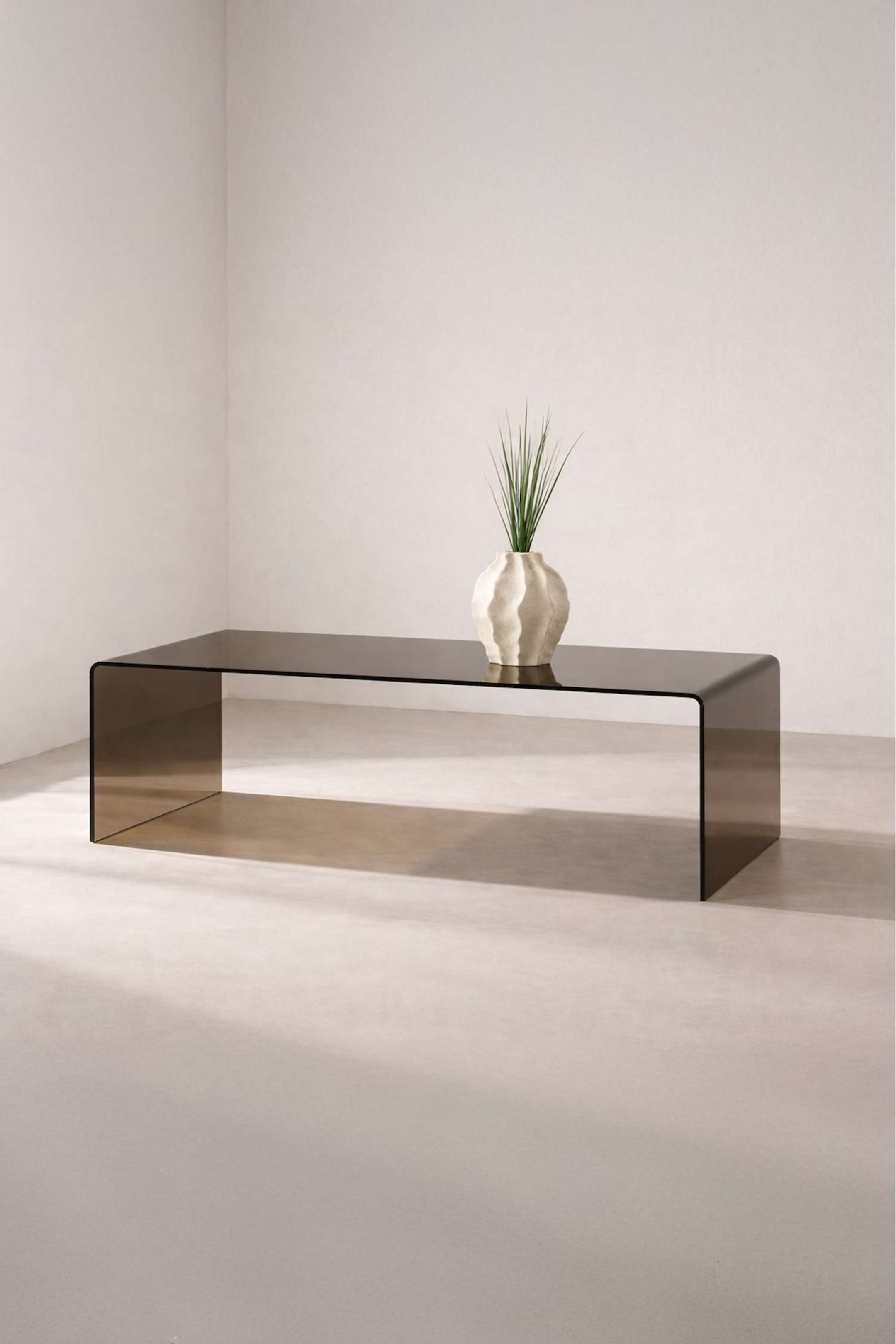 Agrandir l'image: Une table basse en verre fumé présente un plateau rectangulaire et deux pieds légèrement incurvés, formant un U continu, présentée à un léger angle vers la droite. Un vase texturé blanc avec un feuillage vert repose sur sa surface.