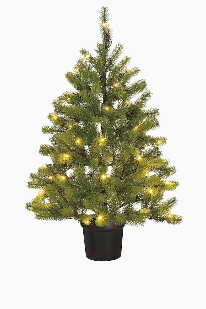 En artificiell julgran står framåtvänd, med täta gröna grenar och många små, varmvita ljus jämnt fördelade. Granen är placerad i en mörk, cylindrisk kruka vid basen. - 1