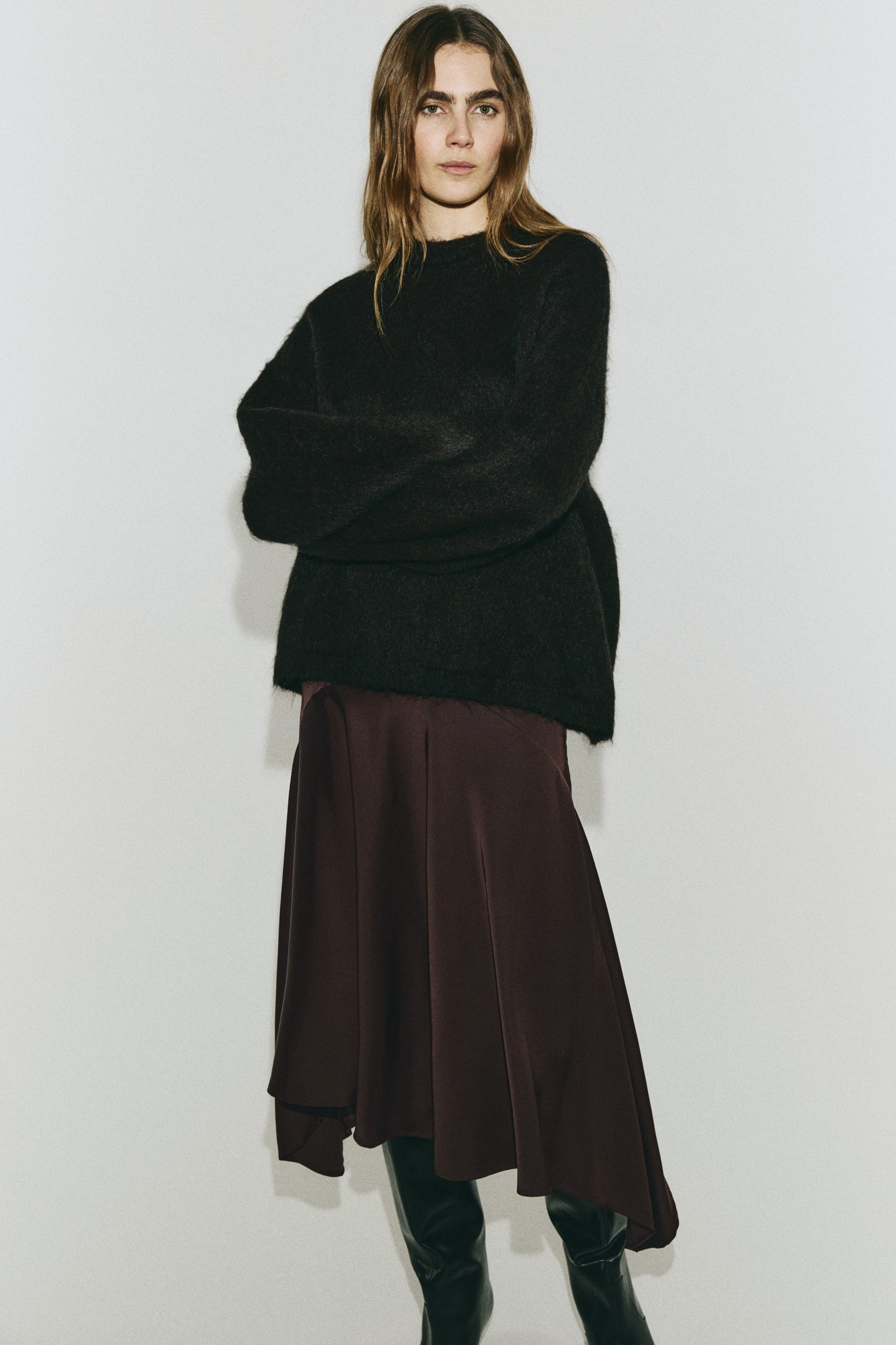 View larger image: A black, fuzzy crewneck sweater features an oversized fit and long sleeves, layered over a dark red midi skirt. The skirt showcases a flowing silhouette with an asymmetrical hemline and a subtle sheen.