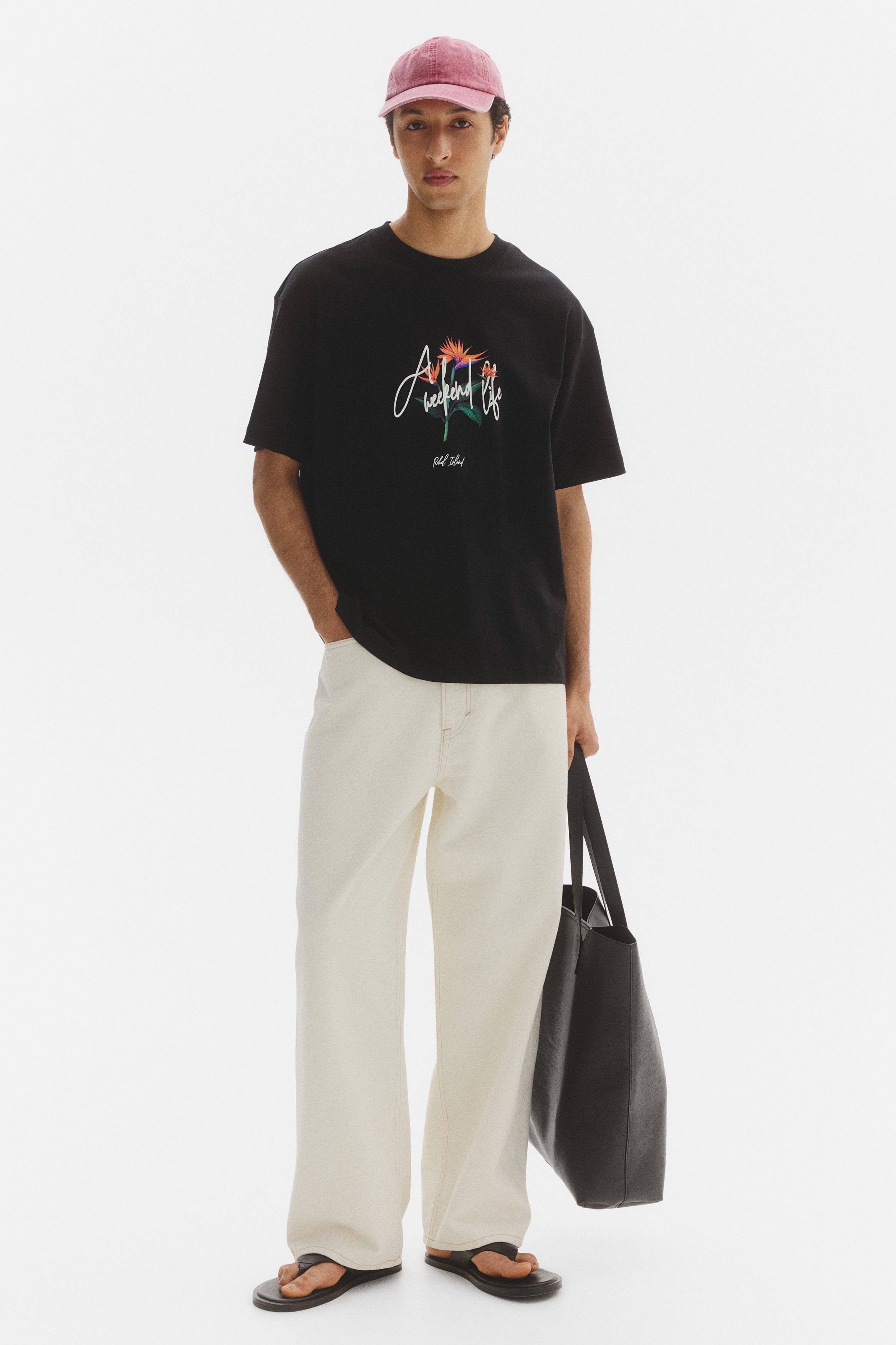 A black t-shirt showcasing a crew neck and short sleeves, viewed from the front. The front displays a graphic print featuring a vibrant bird of paradise flower, accompanied by white script text reading "A weekend life" and smaller script text "Rebel Island" beneath.