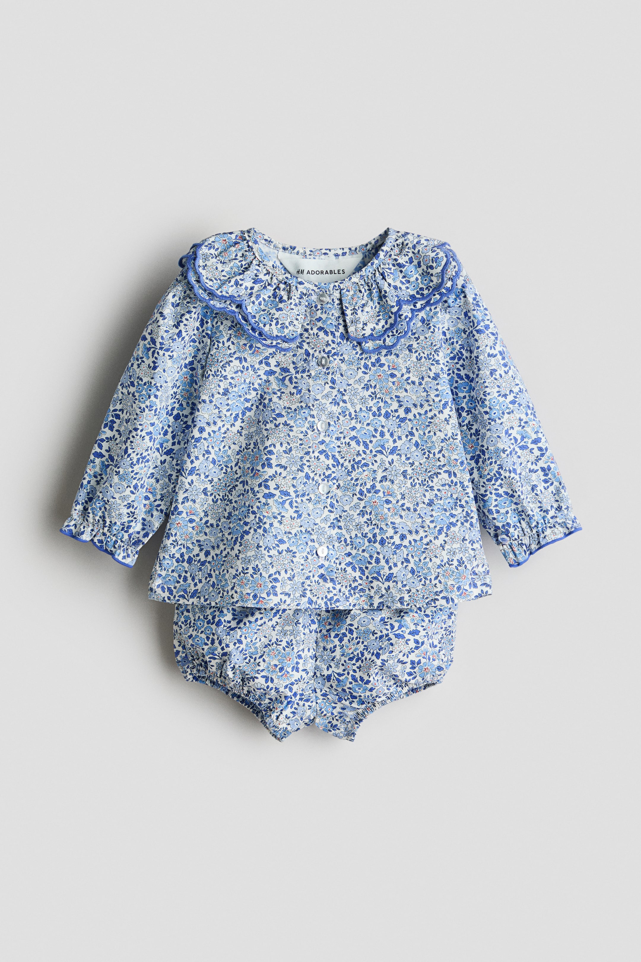 Ampliar la imagen: Un conjunto de blusa y bombachos con estampado floral azul y blanco está presentando su vista frontal. La blusa presenta un cuello doble con borde festoneado y ribete azul, botones al frente y mangas largas con puños fruncidos y elásticos; los bombachos coordinados muestran elastique cubierto en la cintura y las piernas.