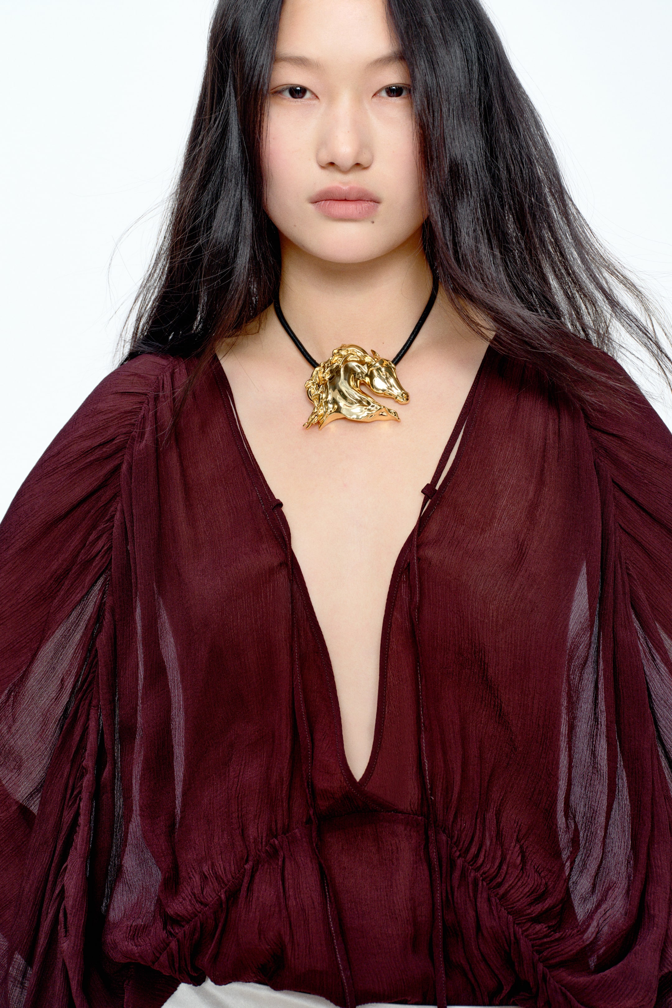 View larger image: A gold-toned necklace with a sculpted horse head pendant facing right, styled on a black cord. The necklace rests against the skin and is visible above a deep V-neck, burgundy sheer top.