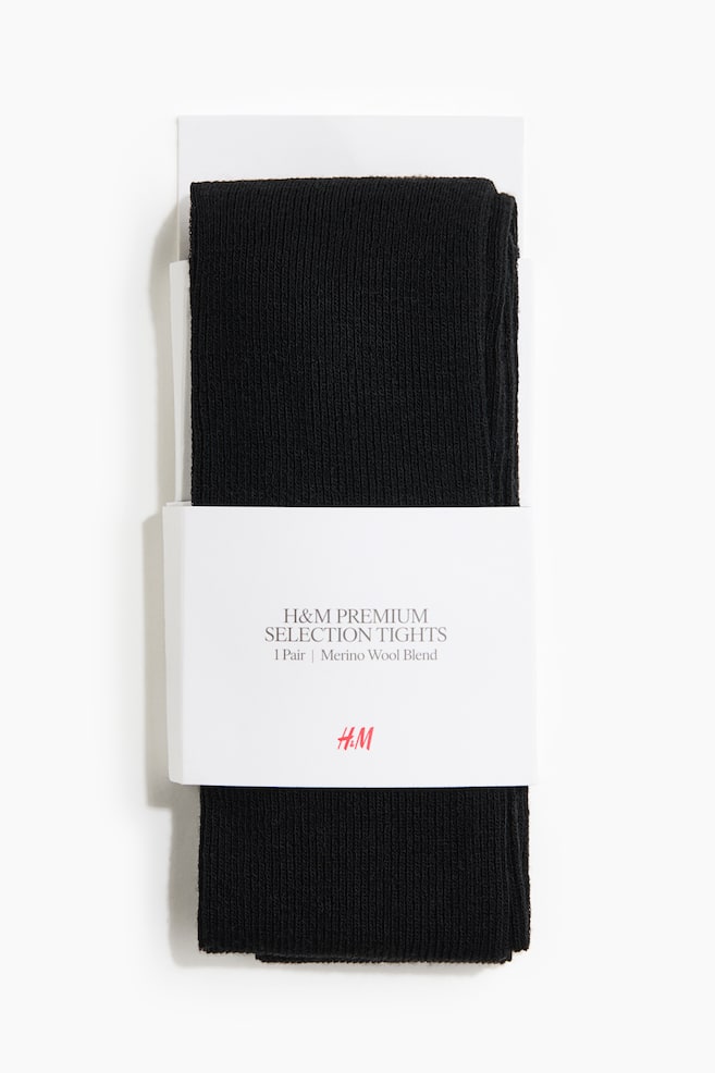 A front view showing black rib-knit tights folded and packaged with a white cardboard backing. A wide white paper band is wrapping around the middle of the tights, displaying the product details and brand logo. - 1