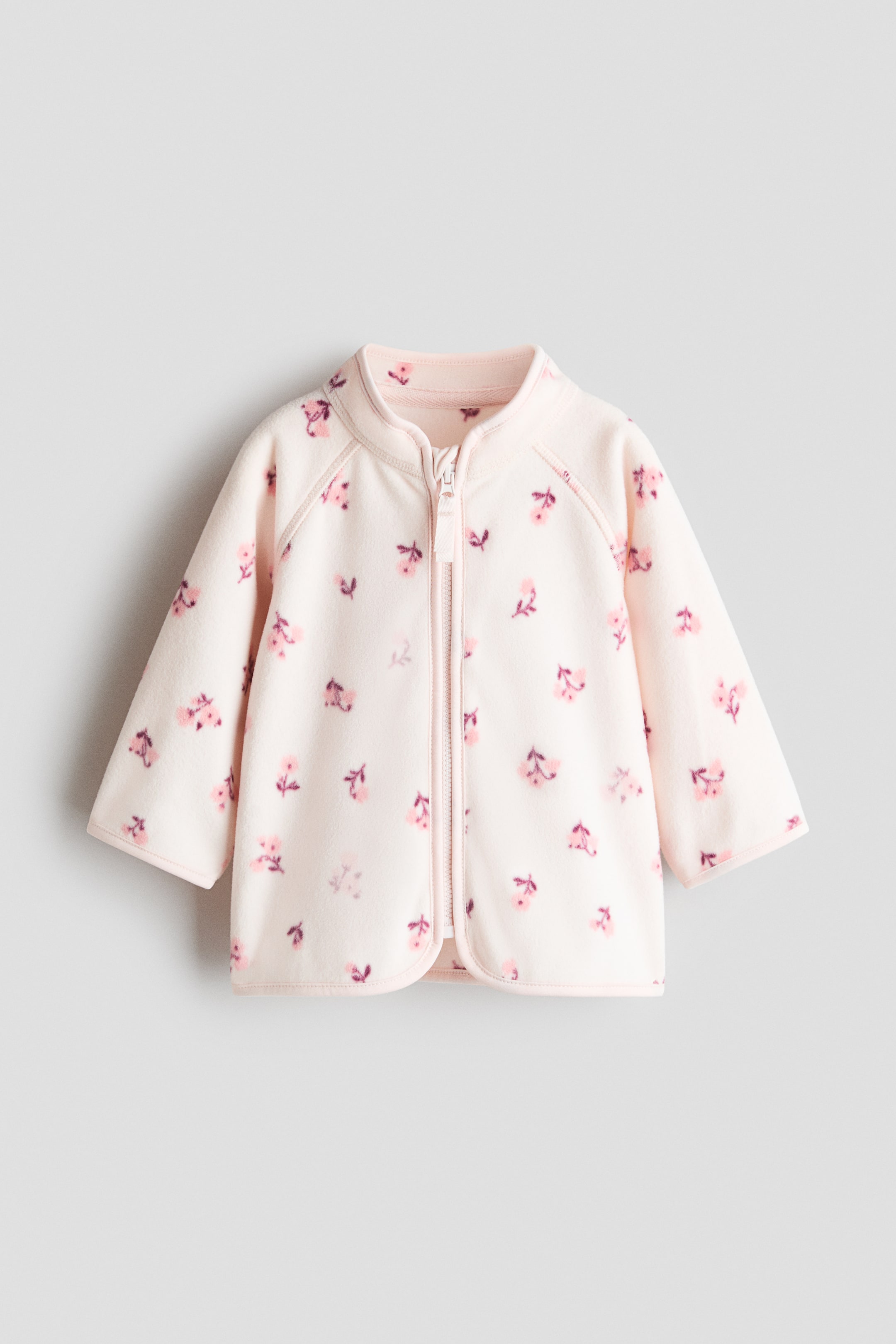 View larger image: The light pink fleece jacket is laid out flat, showcasing a scattered pink floral pattern. It boasts a stand-up collar, a partially open light pink front zipper with an anti-chafe chin guard, long raglan sleeves, and light pink jersey trims at the collar, cuffs, and hem.