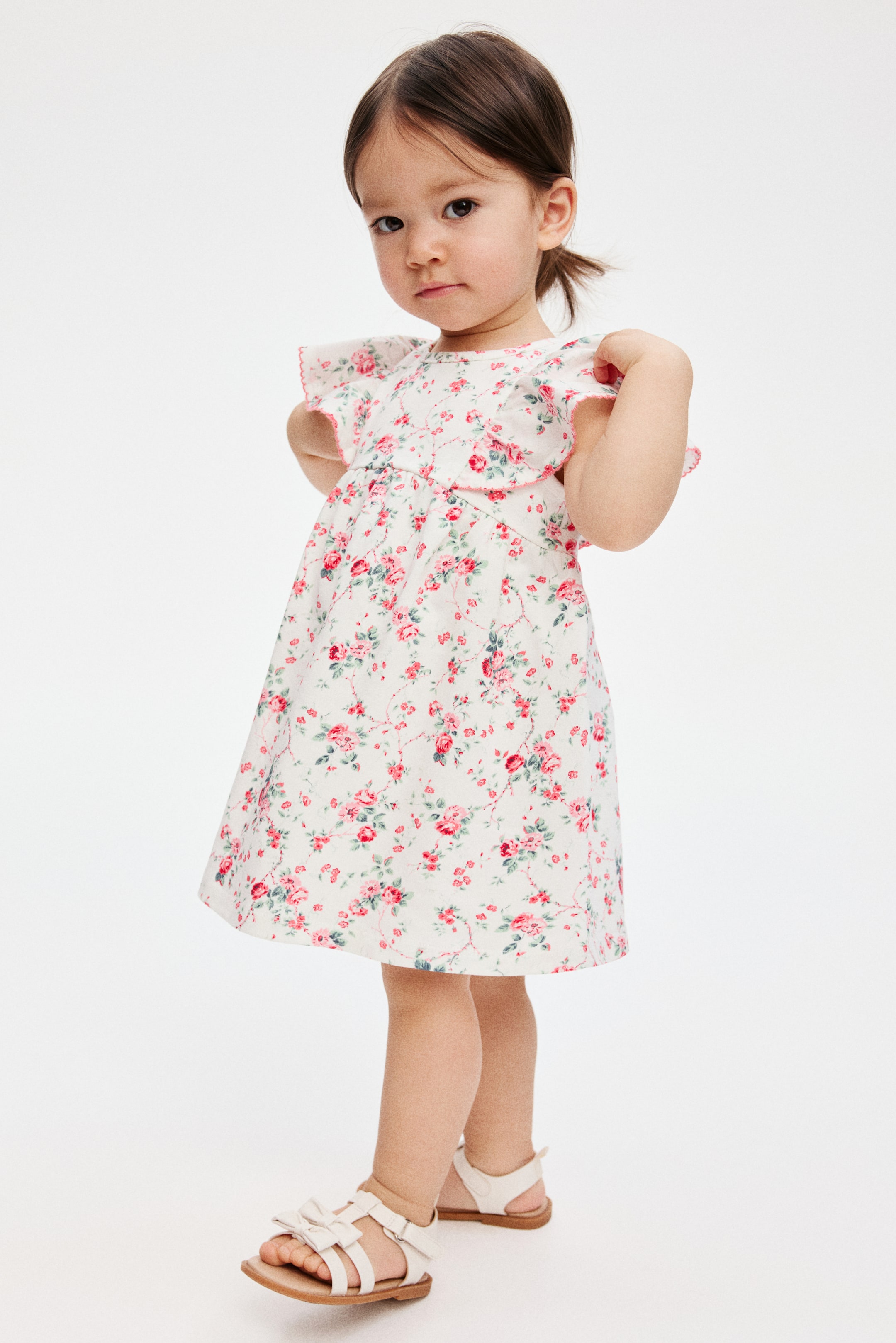 View larger image: "A white dress showcasing a floral print with pink roses and green leaves, accented by pink trim on the rounded neckline and ruffled cap sleeves, and a gathered waistline that creates a flared skirt."