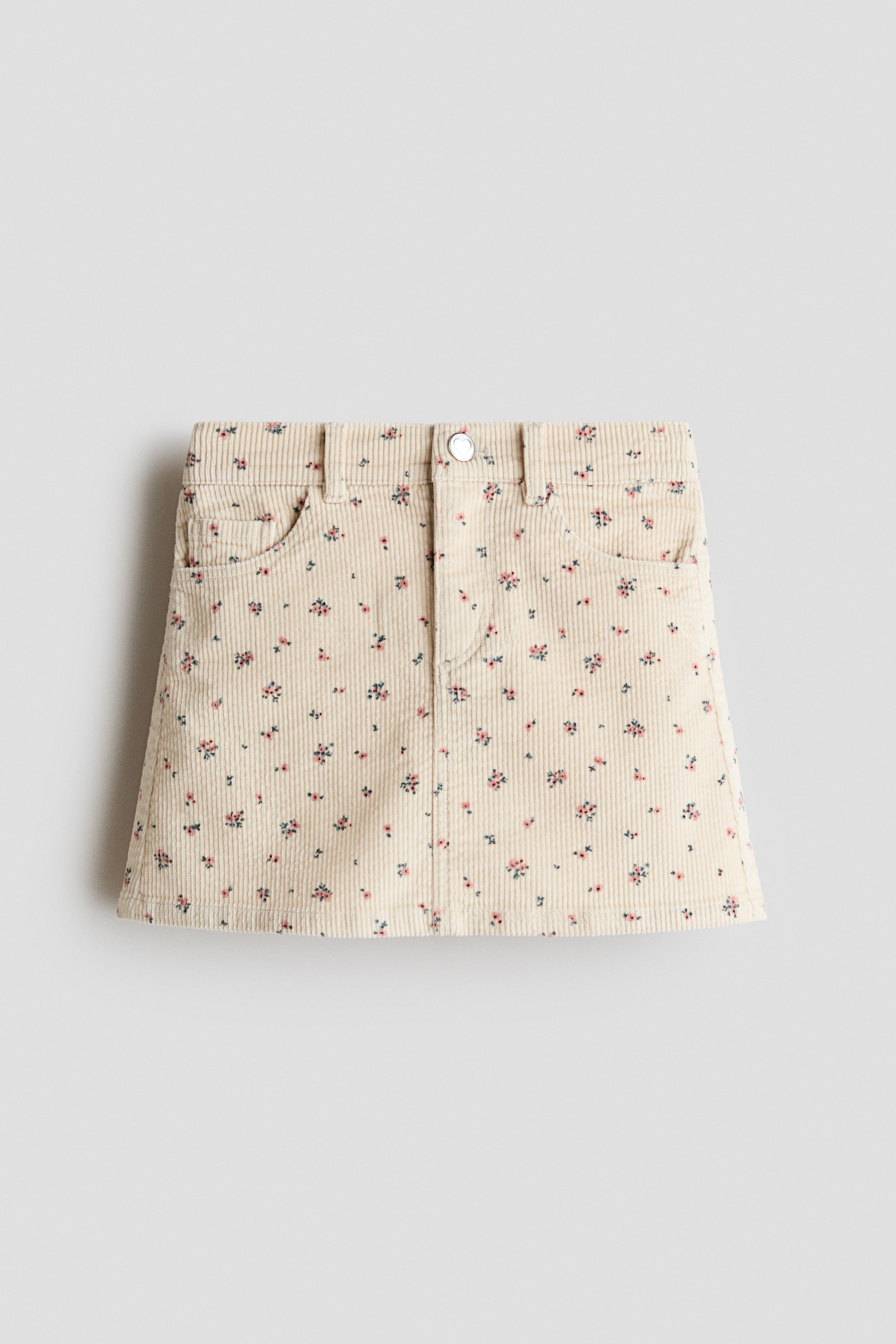 View larger image: A corduroy skirt is shown from the front, featuring beige corduroy fabric with a small floral print. It is designed with a waistband including belt loops, a silver-toned button fastening, and two curved front pockets.