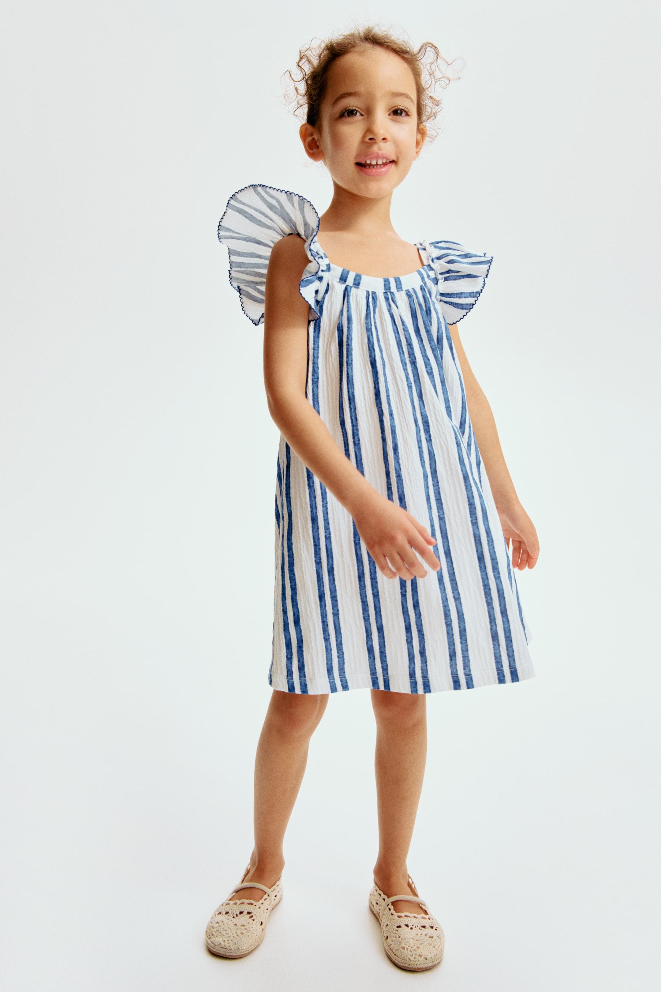 View larger image: A child's blue and white striped dress is showcased from the front, angled slightly to the right. It boasts a crinkled texture with vertical blue stripes, a gathered scoop neckline, and ruffled cap sleeves with blue trim, falling above the knee in a loose fit.