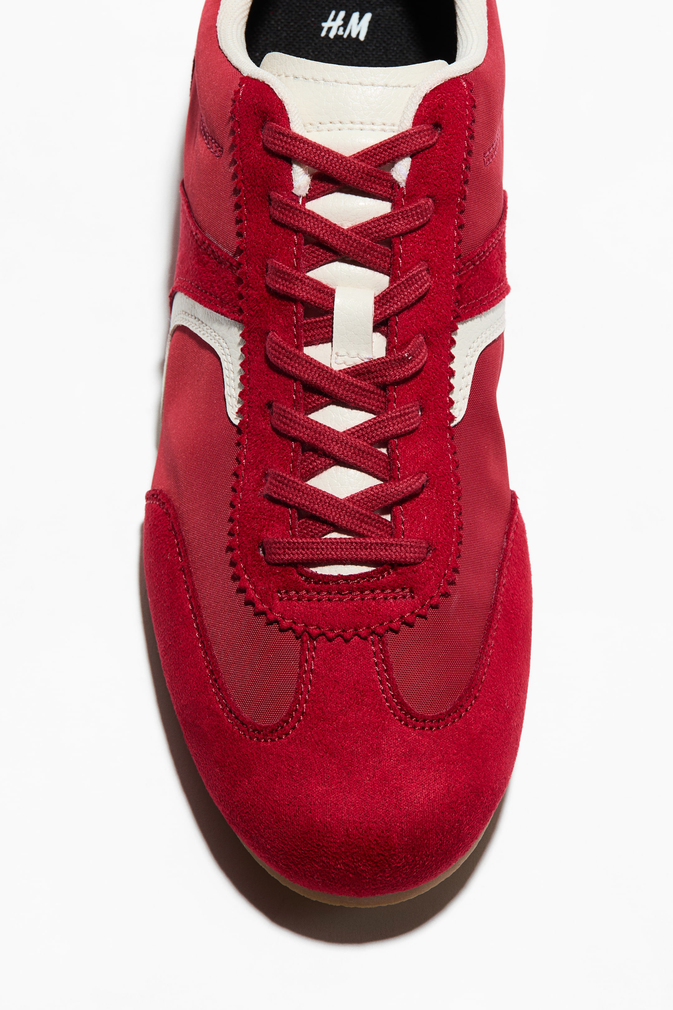 View larger image: A red sneaker is showcased from the front, featuring red laces neatly tied over an off-white tongue. The upper is crafted with red suede and fabric panels, highlighted by off-white stripes on the sides, while the black inner sole displays the H&M logo.