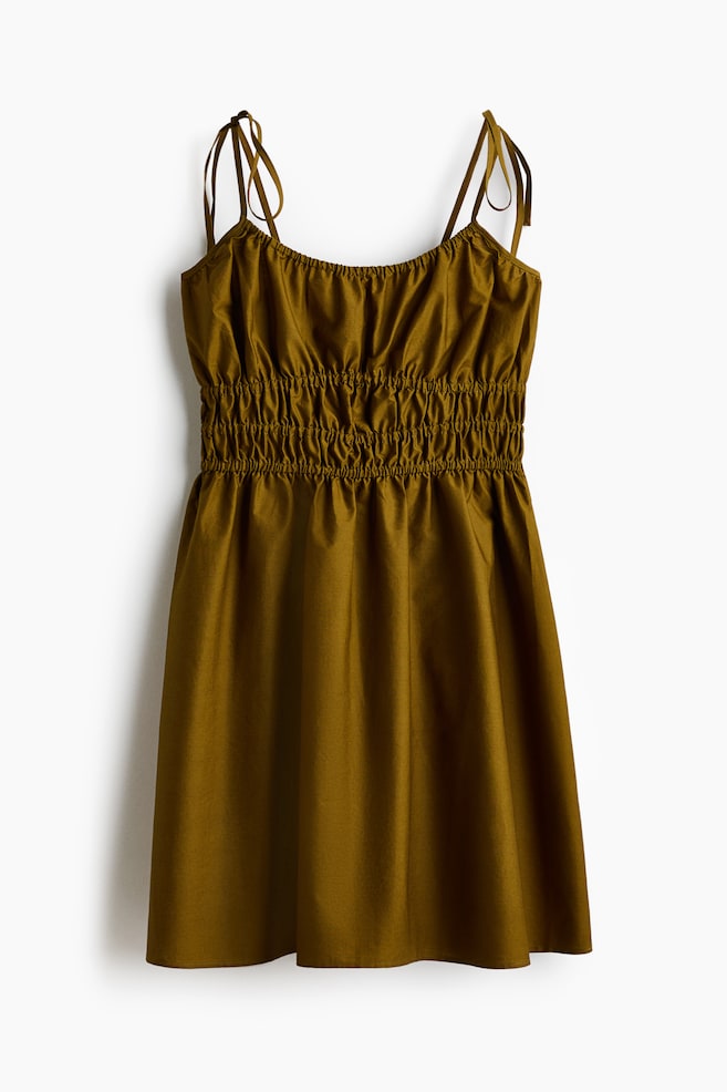 The short dress is displayed front on, featuring a deep, round neckline with covered elastication. It is designed with narrow, tie-top shoulder straps, smocking at the waist, and a flared skirt in an olive green, airy cotton weave. - 1