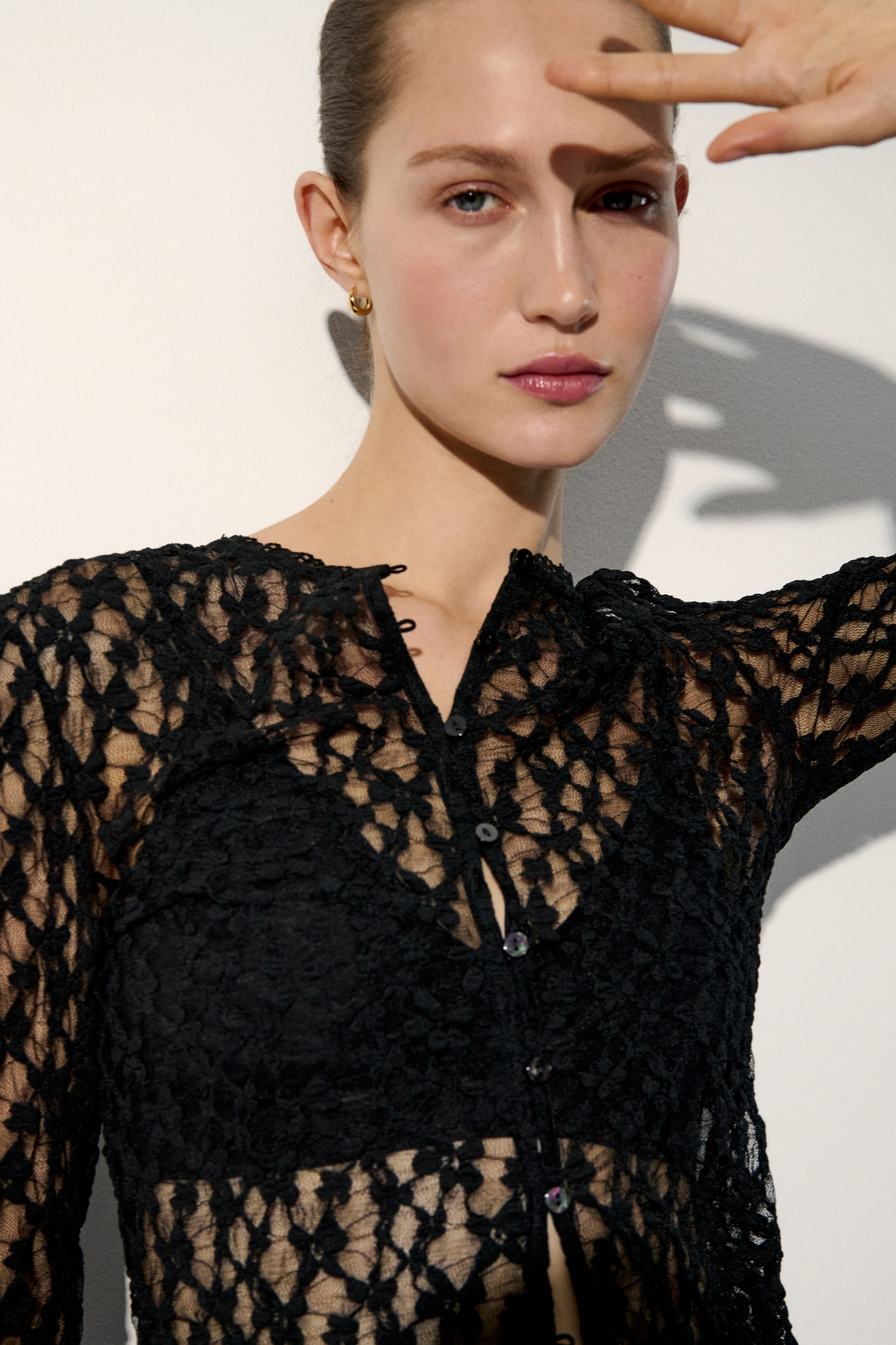 View larger image: A black lace button-front top is displayed from the front, slightly angled to the right, featuring a V-neckline and long sleeves. It showcases a sheer, open-knit lace pattern and is fastened with iridescent buttons, layered over a black bralette.